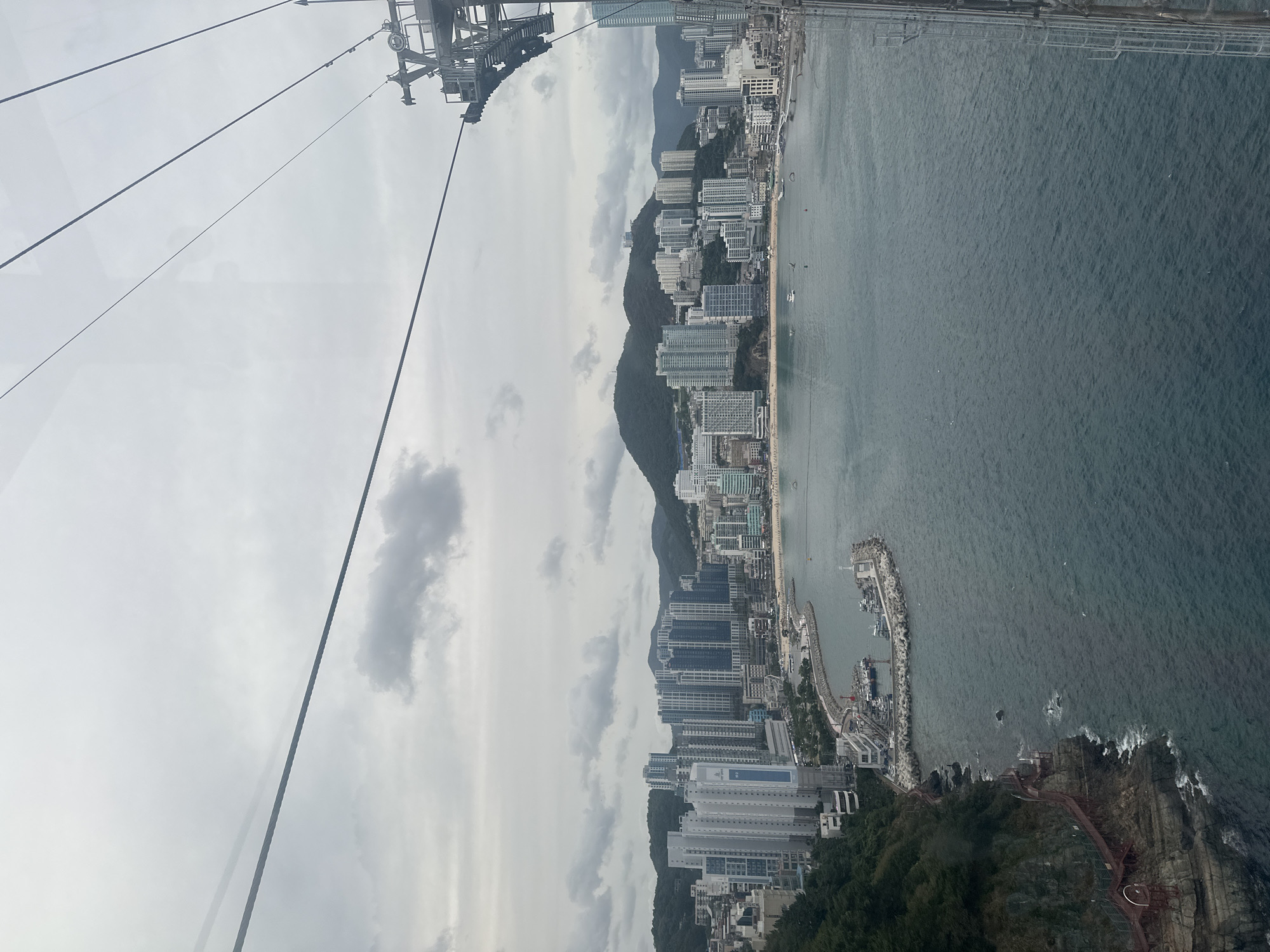 The image, taken from a cable car in Busan, South Korea, offers a panoramic view of the coastal cityscape and the ocean under a cloudy sky. In the foreground, thick steel cables of the cable car crisscross the frame. Below, the dark blue ocean stretches out, dotted with a few small boats and buoys.
The coastline features a dramatic rocky cliff face on the left, adorned with a red wooden walkway snaking its way up its side. At the base of the cliff, a small harbor is visible, with several fishing boats moored within a protective breakwater.
Further inland, a crescent-shaped beach with a few scattered beach umbrellas and people is visible. Behind the beach, a dense urban landscape rises, characterized by numerous high-rise apartment buildings in shades of white, gray, and blue. These buildings form a striking contrast against the backdrop of lush, green mountains that dominate the horizon. The city lights are not yet visible, suggesting it is daytime. The overall impression is one of a bustling coastal metropolis nestled between the sea and verdant hills.