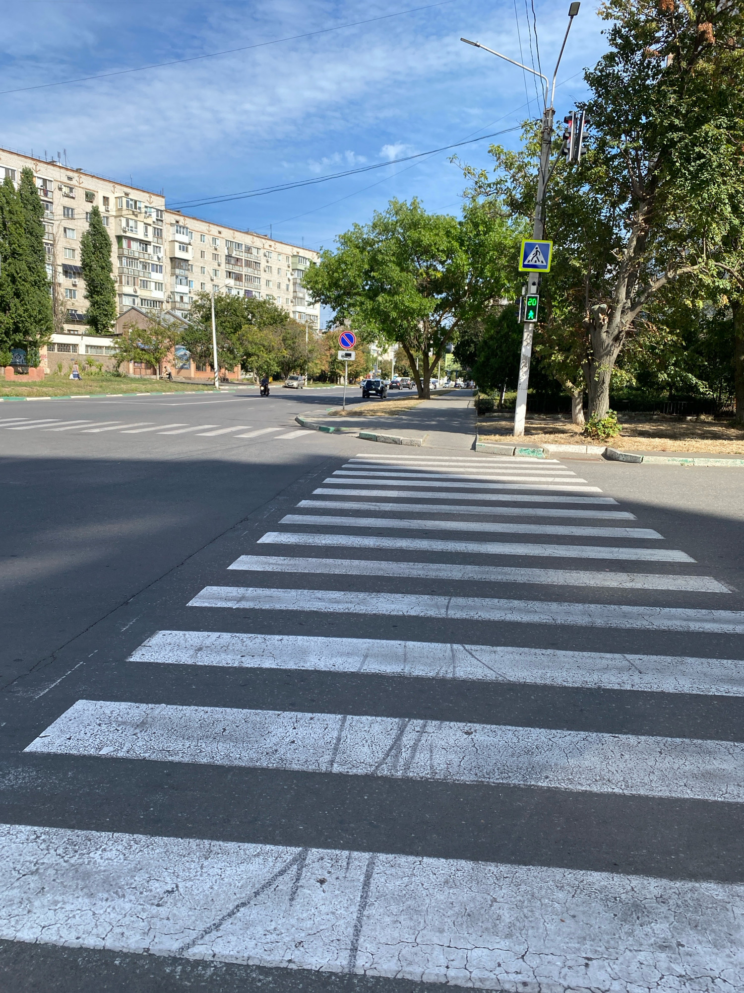The image depicts a crosswalk on a sunny day in Chornomorsk, Ukraine. The crosswalk is in the foreground, with white stripes painted on asphalt. In the background, there is a multi-story apartment building with a light-colored facade and numerous windows. Trees line the street, and a traffic light with a green pedestrian signal is visible. There are a few cars and a person on a scooter on the road in the distance. The sky is blue with scattered clouds.
