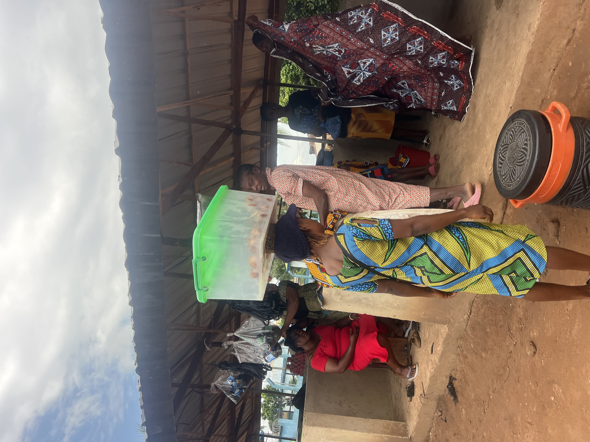 In Nkpor, Nigeria, under a partially open-roofed structure, a group of individuals are gathered. A woman in a patterned dress carries a large, clear green plastic bin filled with red fruits on her head. To her right, another woman in a light-colored, patterned dress stands, while a third woman in a red dress is seated on a stool in the background. A large, patterned fabric is displayed or held by someone to the right. Various items, including what appear to be bags and a dark, orange-topped cylindrical container, are visible on the ground. The setting suggests a market or communal gathering area, with a dirt ground and a rustic overhead shelter.