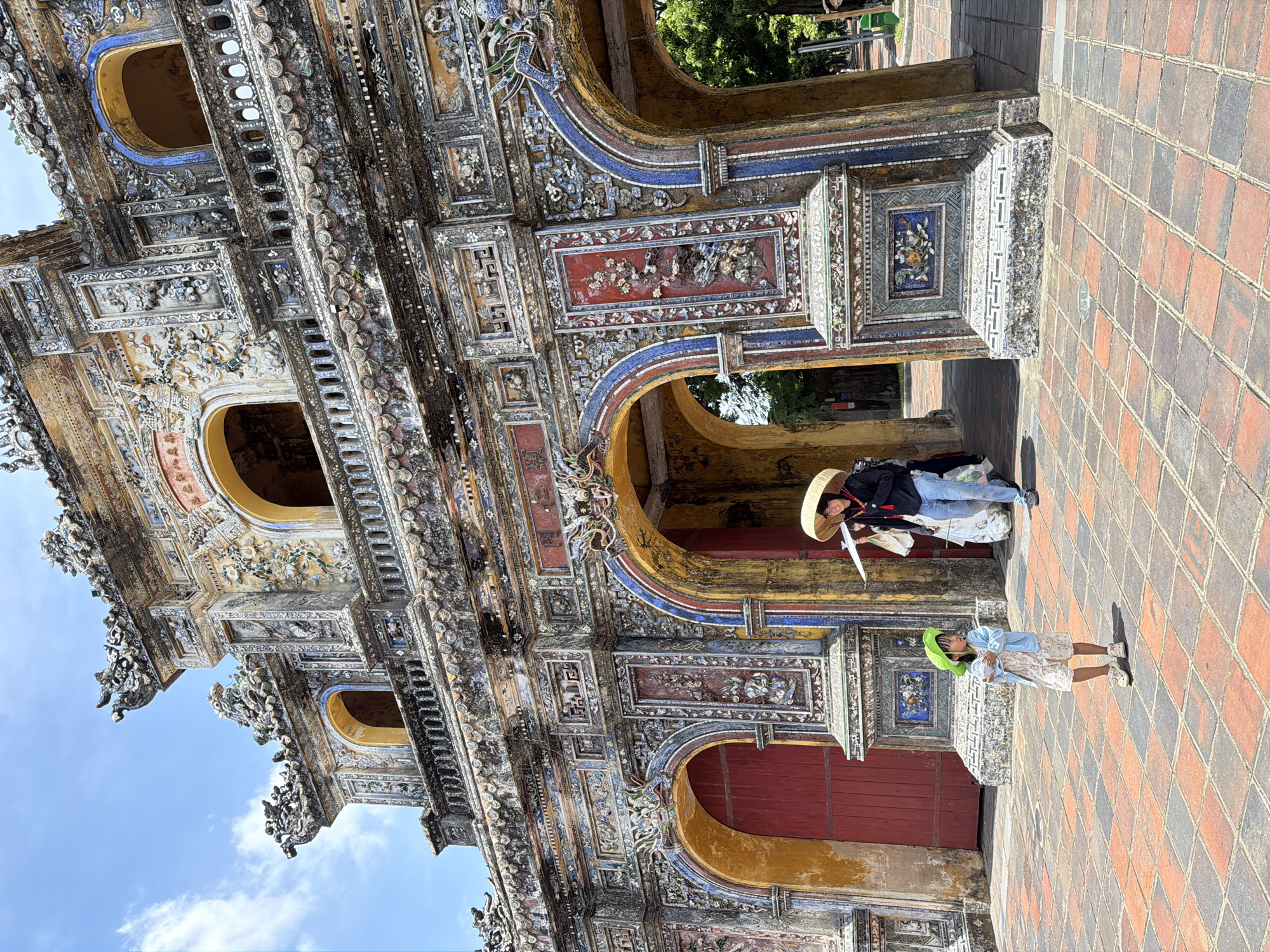 This image depicts the Ngu Phung Gate, part of the Imperial City in Phú Xuân Ward, Vietnam. The ornate, multi-tiered gate features intricate mosaic and stucco decorations, with arched passageways and a central structure. Two people are visible in front of the gate. A young child, wearing a green hat and a light blue jacket over a floral dress, stands in the foreground, holding a white fan. Further back, a woman wearing a conical hat, dark jacket, and jeans walks through one of the arches, carrying a white bag and an umbrella. The ground is paved with orange and grey square tiles. The sky is blue with scattered white clouds.