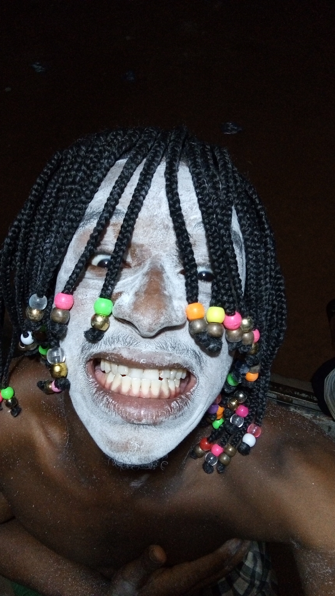 A medium-shot image captures a person with dark skin and a face covered in white paint. The individual has black, braided hair adorned with colorful beads. The person's expression is one of a wide, toothy grin. The environment appears to be indoors, with a dark background. There is not enough information from the image to determine a specific location in the city of Onitsha, Nigeria, or to confirm that this image was taken in Nigeria.
