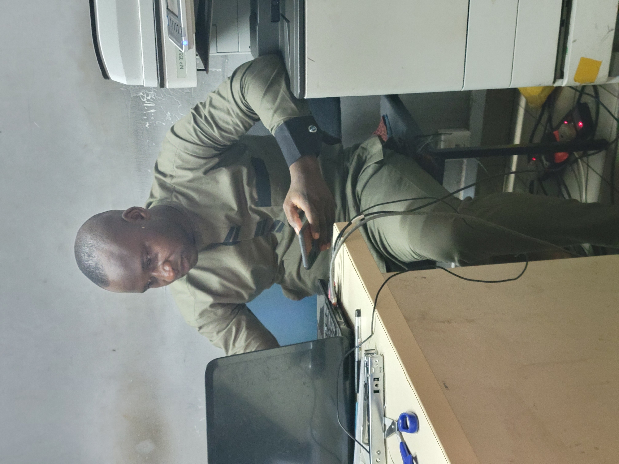 A man is standing in an office in Abuja, Nigeria, near a desk and a photocopier. He is dark-skinned, bald, and is wearing a long-sleeved, olive-green uniform. He is holding a black cell phone in his hand, operating it in proximity to a laptop. The desk is cluttered with office supplies, including a pair of blue scissors and a stapler. The photocopier is a large, white multifunction machine. The wall is painted white. Wires are tangled behind the desk and around the photocopier. A power strip with red indicator lights is visible.
