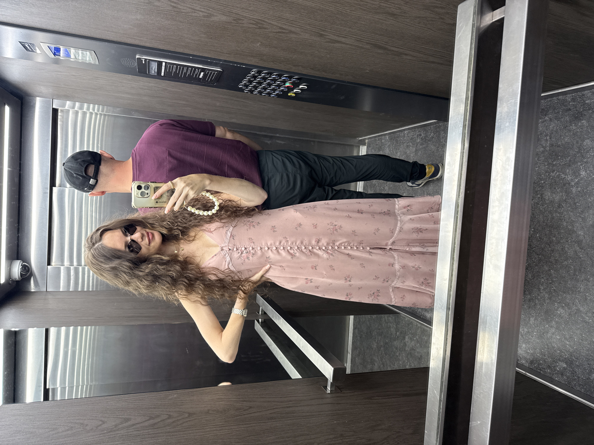 An interior shot taken in a wood-paneled elevator in Moscow, Russia, shows a young woman and a man. The woman, facing the camera, wears a pink floral dress, sunglasses, and a pearl necklace. She holds a smartphone, taking a selfie. The man, facing away from the camera, wears a maroon t-shirt and black pants. An elevator control panel is visible on the right wall.