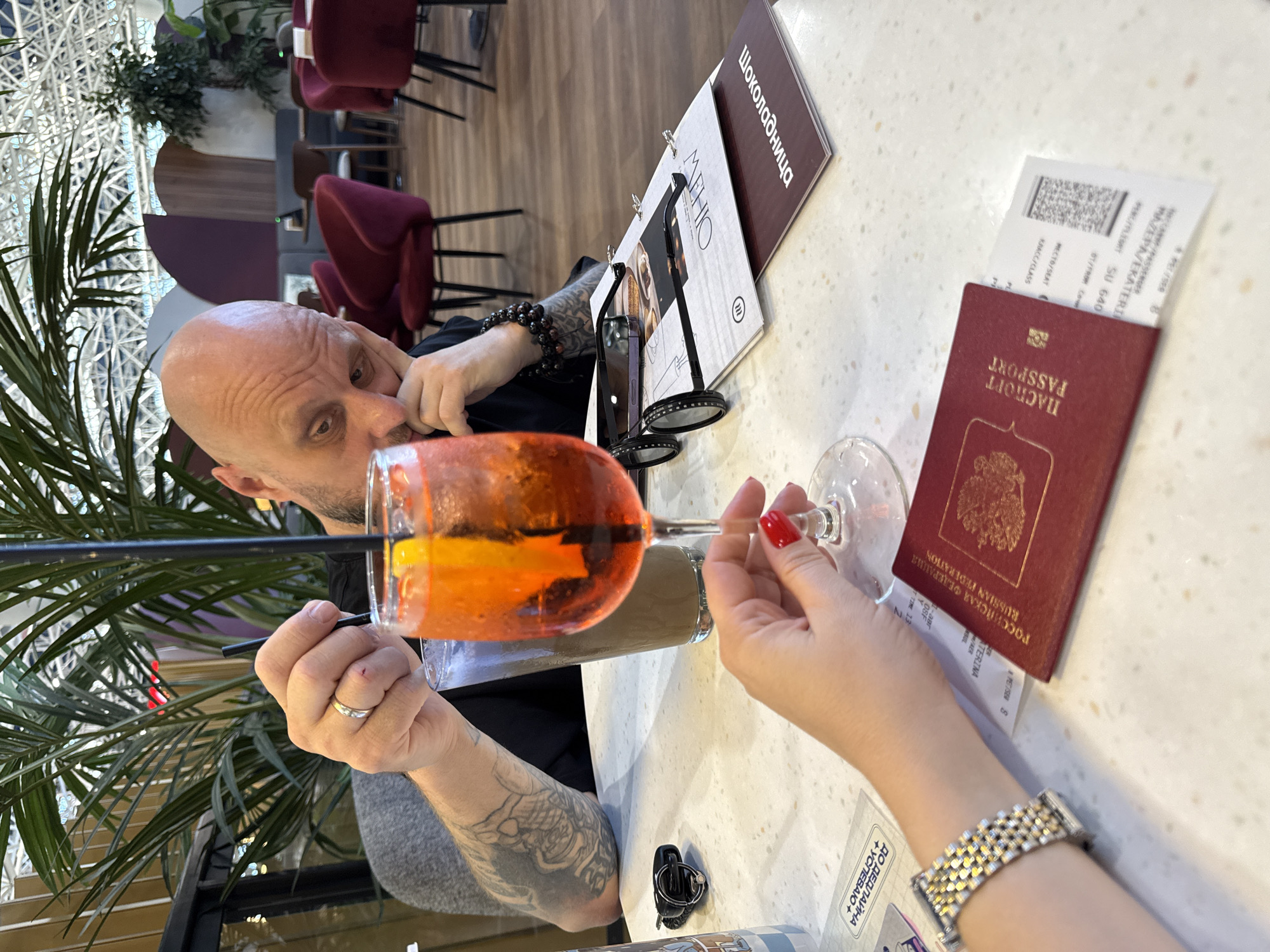 The image shows a close-up view of a table in what appears to be a cafe or restaurant. In the foreground, a woman's hand with red nail polish is holding a wine glass filled with an orange beverage garnished with an orange slice and ice. To the left of the glass, a man's arm with tattoos and a wedding ring is visible, holding a black straw in a glass of iced coffee. On the table, a Russian Federation passport and a boarding pass are partially visible. There are also eyeglasses and a menu laid out on the table. In the background, there are plush red chairs and plants, suggesting an indoor seating area. The overall lighting is soft, and the focus is on the foreground elements.
The provided context indicates the location is Sochi, Russia. Based on the visible passport and boarding pass, the scene likely depicts individuals preparing for or having just completed travel, possibly related to an event or vacation in Sochi. The presence of a menu and drinks suggests a dining establishment.