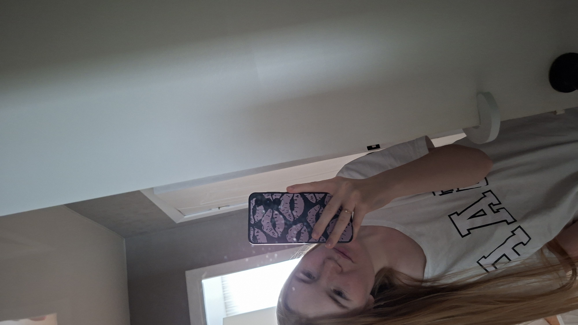 A young woman with long blonde hair takes a mirror selfie in what appears to be a residential bathroom. She is wearing a white t-shirt with large black block letters on it. She is holding a smartphone with a black case decorated with purple lips. Her right hand, wearing a ring, is visible holding the phone. The background shows a tiled wall, a doorframe, and a white door with a handle. The lighting suggests it is daytime, with natural light coming from an unseen window or opening. There are no discernible elements that would definitively place this scene in Chaikovsky, Russia.