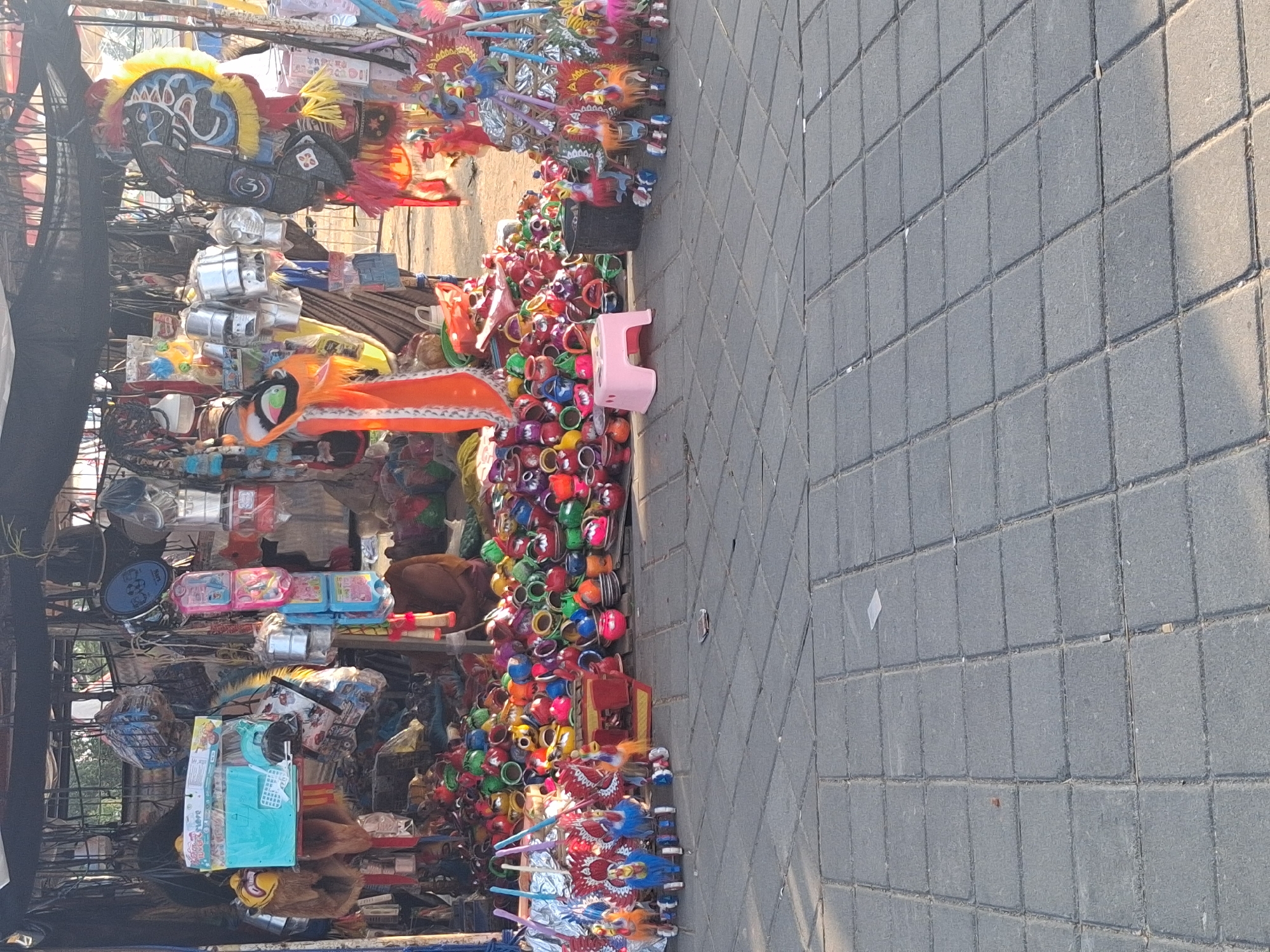 A street stall in Surakarta, Indonesia displays a variety of colorful toys and traditional crafts. The stall is covered by a dark tarp, and behind the merchandise, a person is partially visible, seated and wearing a brown garment. The foreground is paved with gray stone tiles. The merchandise includes numerous small, brightly painted clay pots, wooden horse toys on wheels with colorful manes and tails, and some larger decorative items hanging from the stall. Among the items are also what appear to be lunch boxes and small plastic stools. The scene suggests a lively marketplace environment.