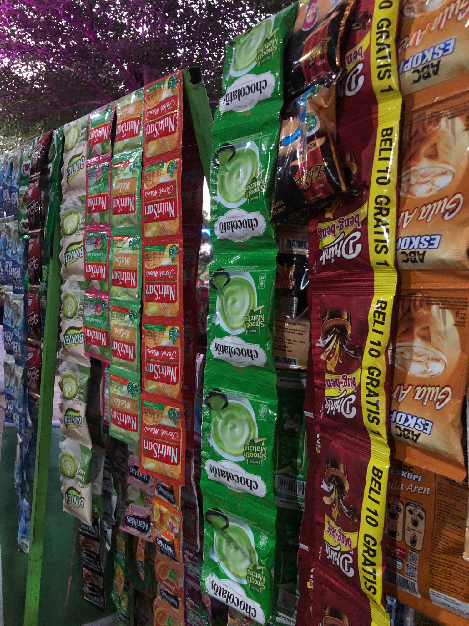This image depicts a stall in Sukoanyar, Indonesia, displaying an assortment of packaged food and beverage items. The stall features rows of colorful sachets hanging from a green frame. These sachets appear to be for powdered drinks, snacks, and possibly instant coffee, with brands like "NutriSari," "Bonten," and "Drink beng-beng" visible. Some sachets have promotional text such as "GRATIS 1" (Free 1) and "BELI 10 GRATIS 1" (Buy 10 Get 1 Free). The background shows a blurred tree with purple foliage and a hint of sky, suggesting an outdoor setting. The overall scene is a typical street-side vendor display focused on convenience items.
