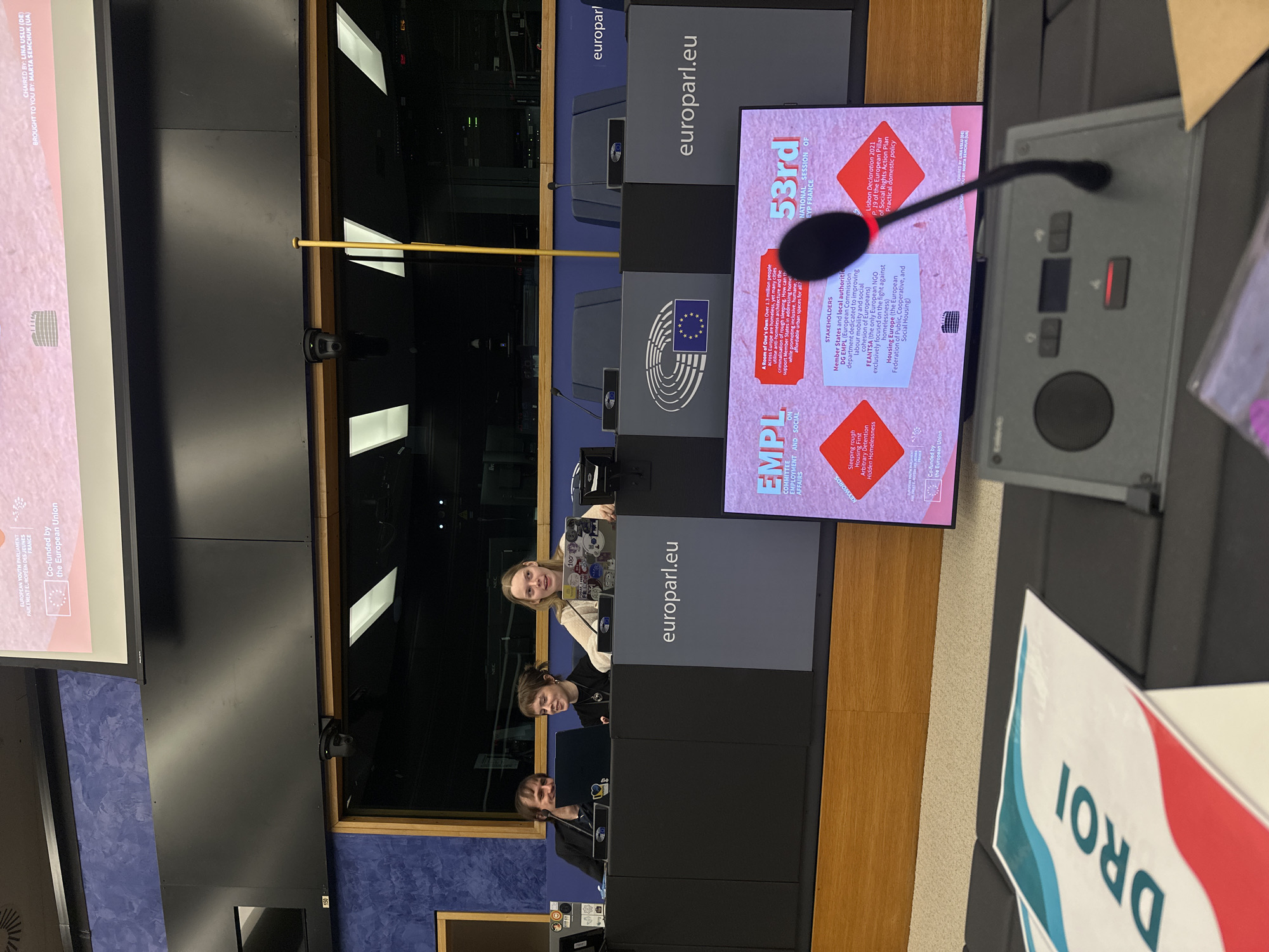 The image shows an interior view of what appears to be a conference or meeting room, likely within the European Parliament in Strasbourg, France. Three people are seated at desks behind a barrier with "europarl.eu" printed on it. A sign reading "DROI" is partially visible on the right side of the image. A screen displays the "53rd National Session of EYP France" with information related to employment, social affairs, and homelessness. A microphone and control panel sit in the foreground. The overall environment seems formal, suggesting a professional setting for discussions or presentations.