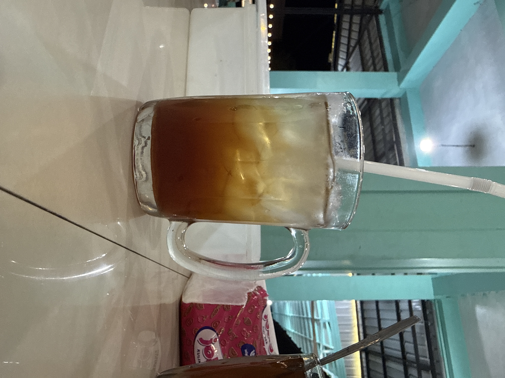 A glass of iced tea sits on a tiled surface in front of a white container. The drink, with ice cubes and a straw, is dark brown at the bottom and lighter at the top, suggesting it is iced tea. To the right, another glass contains a dark liquid, likely coffee or tea. A pink and white tissue packet with a logo is visible between the glasses. The background features turquoise pillars and a roof structure, suggesting an outdoor or semi-outdoor setting, possibly a food court or restaurant in Grong-Grong, Indonesia. The lighting is dim, with a single bulb visible in the distance, and the overall atmosphere is casual.