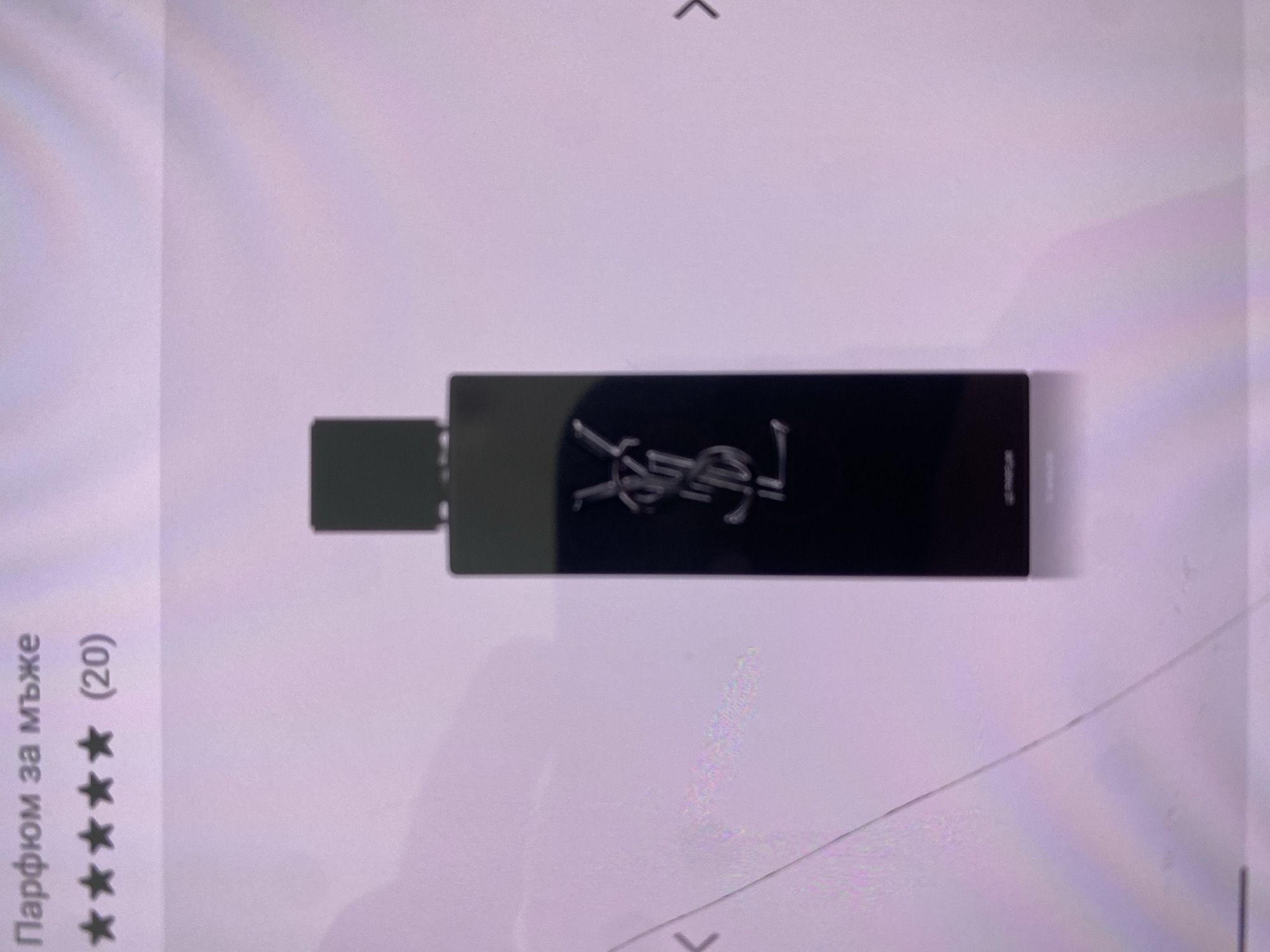 The image shows a bottle of Yves Saint Laurent perfume for men. The bottle is rectangular, black, and has the YSL logo in silver. To the left of the bottle are the words "Парфюм за мъже" (Perfume for men) followed by five stars and "(20)". The background is a light purple or white color. The location is not discernible from the image.