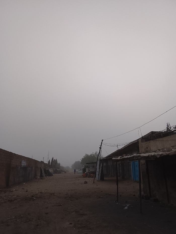 A desolate dirt road stretches into a dense fog, flanked by worn buildings. A lone figure walks in the distance, barely visible through the heavy haze. To the left, a brick wall stands alongside debris. On the right, dilapidated structures with decaying canopies line the street. Power lines sag above the scene, contributing to the atmosphere of decay and isolation. The overall impression is one of neglect and stillness, obscured by the pervasive fog.