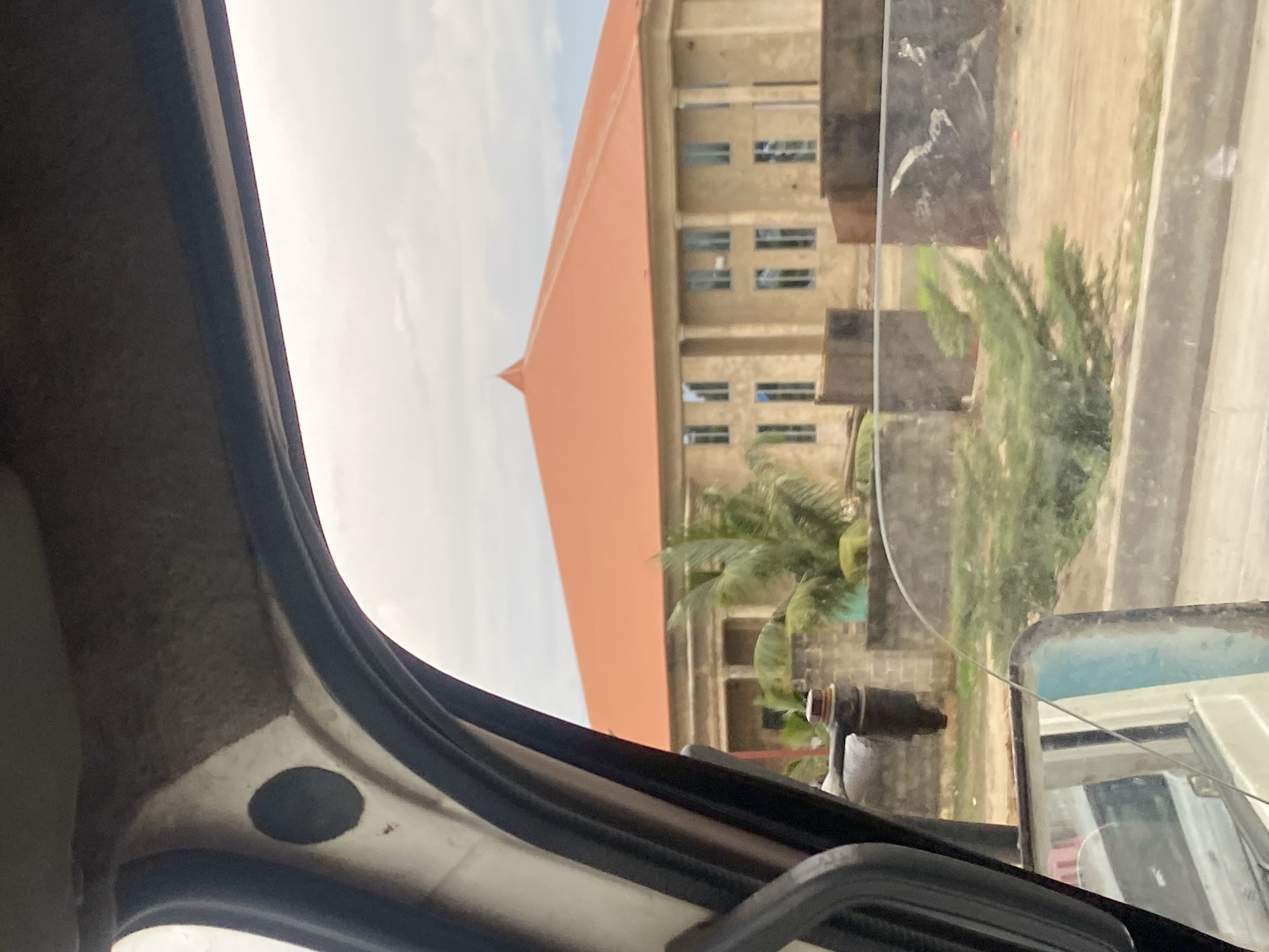 The image is taken from inside a vehicle, looking out through the windshield and side window. In the foreground, the interior of the vehicle is visible, including a part of the dashboard, a side mirror, and the car's A-pillar. The exterior view shows a large building with a distinctive orange, conical roof. The building appears to be constructed of light-colored brick or stucco and features a colonnade along its facade, with several windows visible. In front of the building, there are some trees, including a palm tree, and a low brick wall. The ground is covered in dirt and some sparse vegetation. The sky is overcast. The scene is likely located in Ikorodu, Nigeria, given the context provided.
