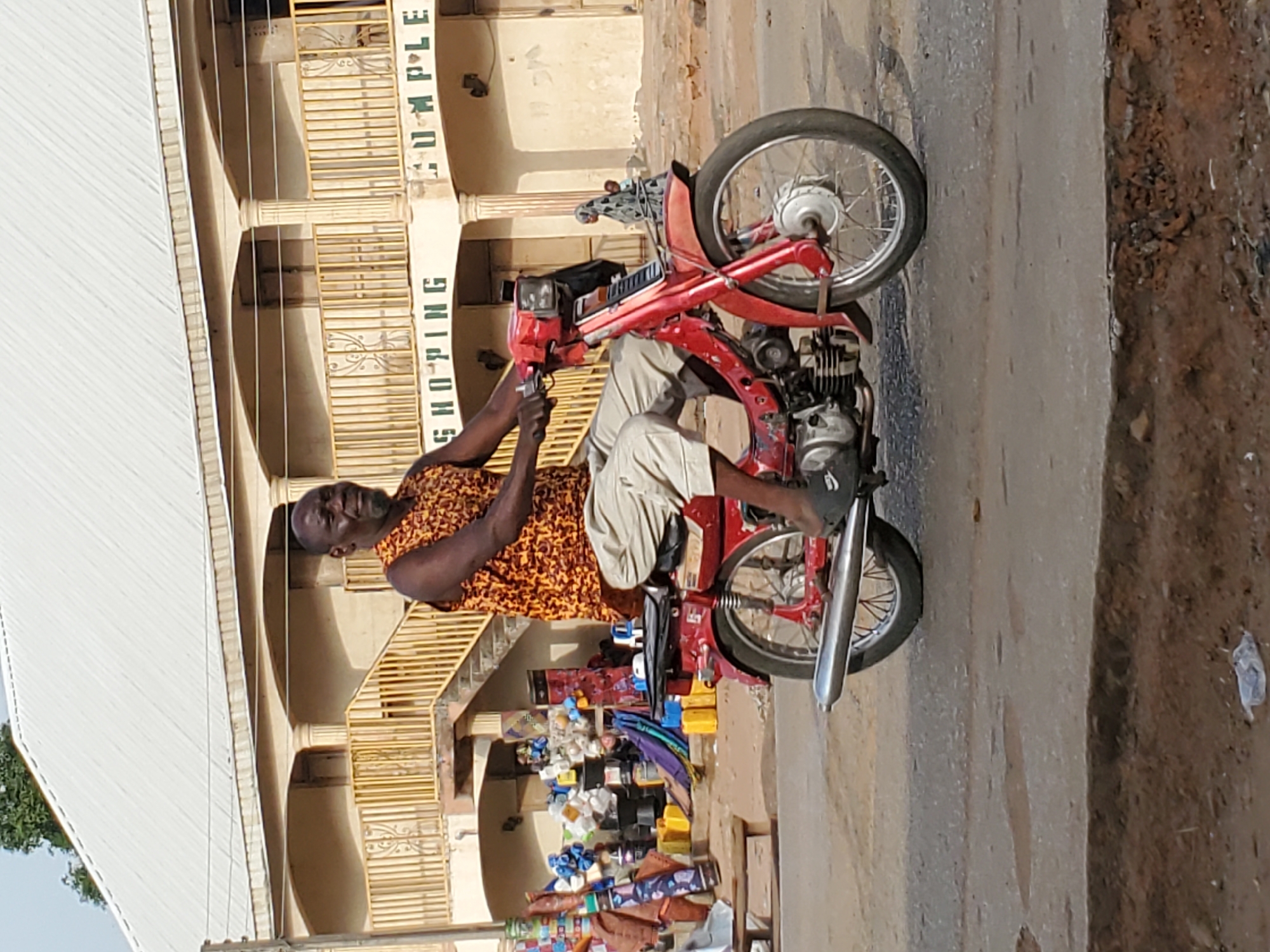 This photograph, taken in Mararaba, Nigeria, depicts a man seated on a red motorcycle during the day. The man, with dark skin, wears a vibrant orange and brown patterned sleeveless shirt and light-colored shorts, along with sandals. He is positioned sideways on the stationary motorcycle, looking towards the viewer with a relaxed, slightly smiling expression, his hands resting near the handlebars.
The motorcycle is a small, red, moped-like vehicle with visible engine components and a silver exhaust pipe. It is parked on a paved road, which transitions into a darker, unpaved dirt area to the right.
In the background, a multi-story, light yellow or cream-colored building with arches and columns is visible. Green letters painted vertically on the building clearly spell out "SHOPPING COMPLEX." Below this text, on the ground level, various merchandise items are displayed, indicating a market or small shops. These items include plastic containers, fabrics, and other colorful goods. The bright, direct sunlight suggests it is daytime, casting clear shadows.