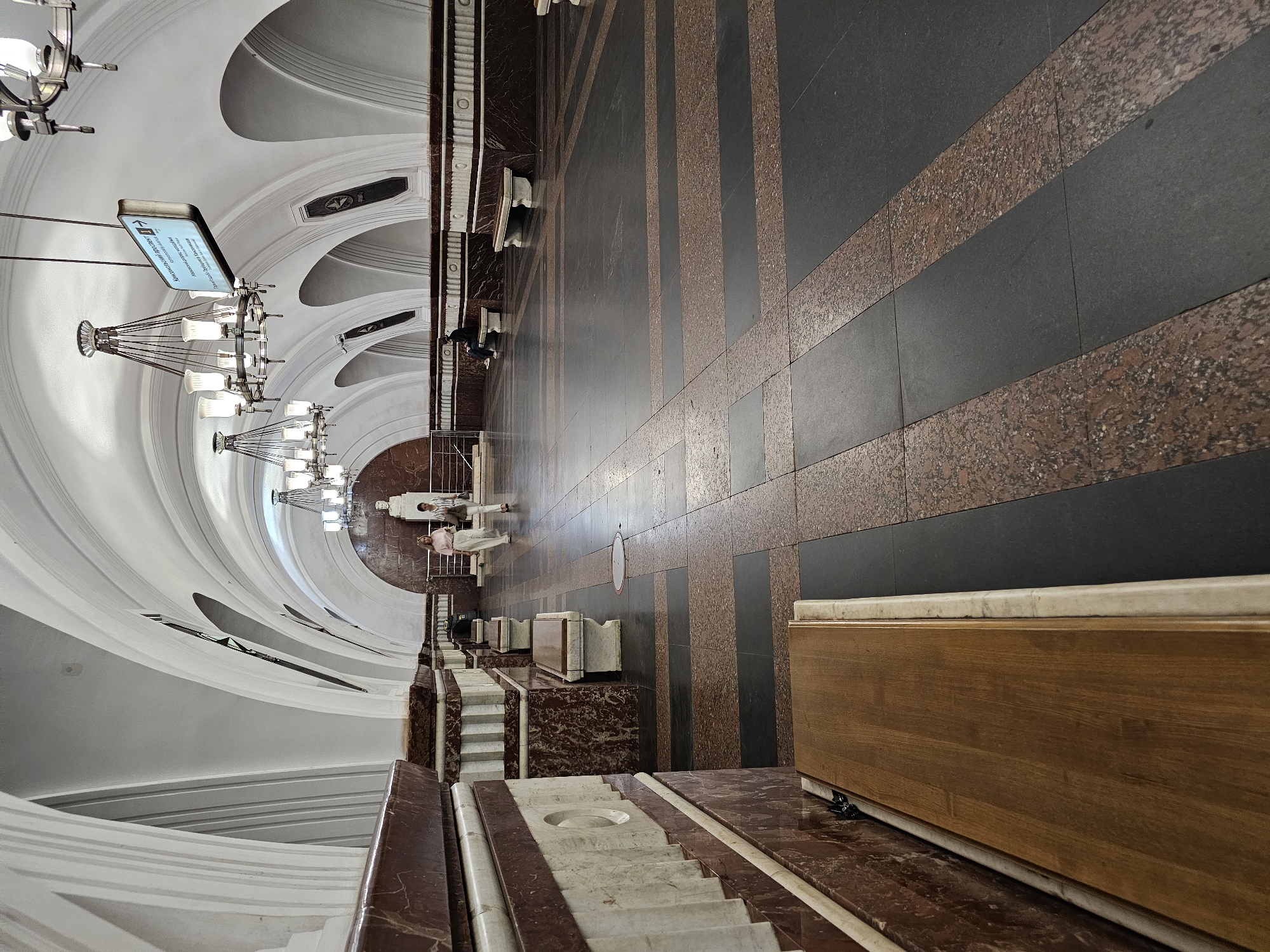 The image captures the interior of the Komsomolskaya metro station in Moscow, Russia. The scene is shot from a high vantage point, looking down the length of the platform. The architectural design is ornate, featuring a high, arched ceiling painted in white, adorned with intricate molding and lighting fixtures. Large chandeliers hang from the ceiling, providing illumination.
The floor is covered with a patterned surface of alternating stripes of black and reddish-brown material. Along one side of the platform, there are benches made of marble and wood, where people are seated. A few people are standing and walking on the platform.
The overall environment suggests a grand and well-maintained space, characteristic of Moscow's metro stations.