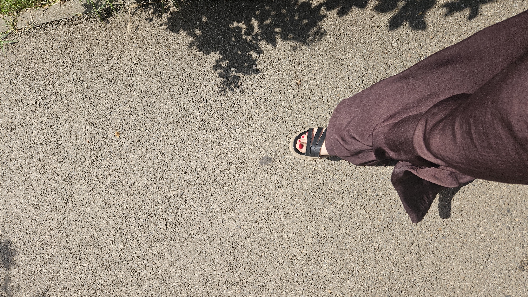 A person's feet are shown walking on an asphalt path in Krasnodar, Russia, on a sunny day. The person is wearing dark brown pants and black sandals with red painted toenails. Shadows of tree branches are cast on the asphalt. To the right of the path, there is a strip of grass and a concrete border. The scene depicts a leisurely walk in an outdoor urban setting.
