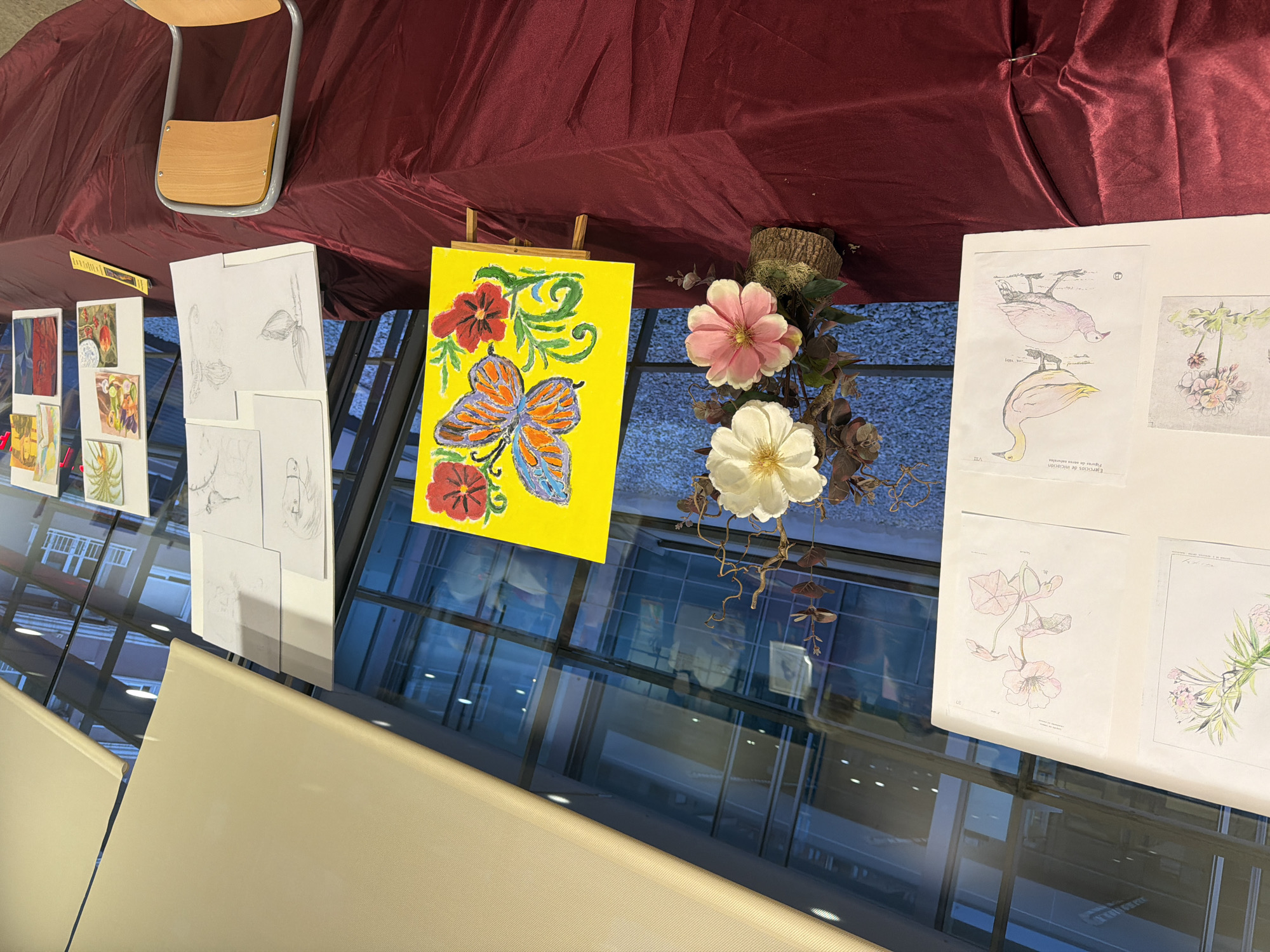 The image depicts an art exhibition displayed on a table covered with a dark red tablecloth. The exhibition features various artworks, including sketches of plants and animals, and a colorful painting of a butterfly on a yellow background, propped on an easel. A floral arrangement with artificial white and pink flowers is placed to the left of the butterfly painting. Behind the artworks, large windows reveal reflections of people and the interior of what appears to be a public building or gallery. The setting suggests a cultural event, possibly an art class or school exhibition, in a contemporary indoor space with ample natural light. The Elche/Elx city context is not explicitly evident from the visual elements within the image itself.