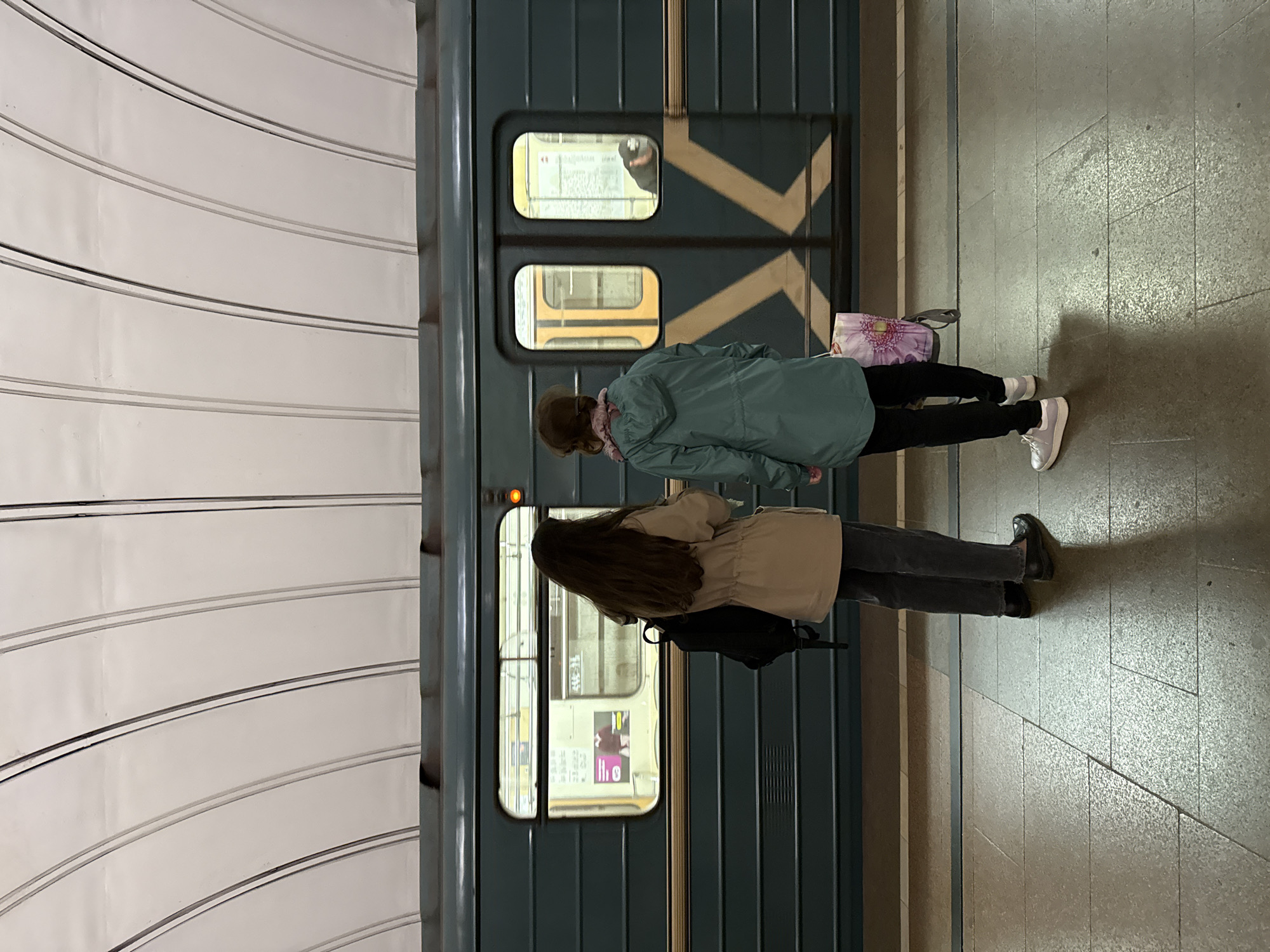 Two young women stand on a metro platform in Moscow, Russia, waiting for a train. The woman on the left, with long dark hair, wears a beige jacket and dark pants, carrying a black backpack. The woman on the right, with shorter brown hair, is dressed in a teal hooded jacket and black pants, holding a patterned tote bag. A teal metro train is stopped at the platform, its doors open. The platform is made of textured stone tiles, with a yellow line indicating the safety edge. The ceiling of the station is a ribbed, curved structure with white panels. The scene is captured from behind the women, facing the train and the platform.