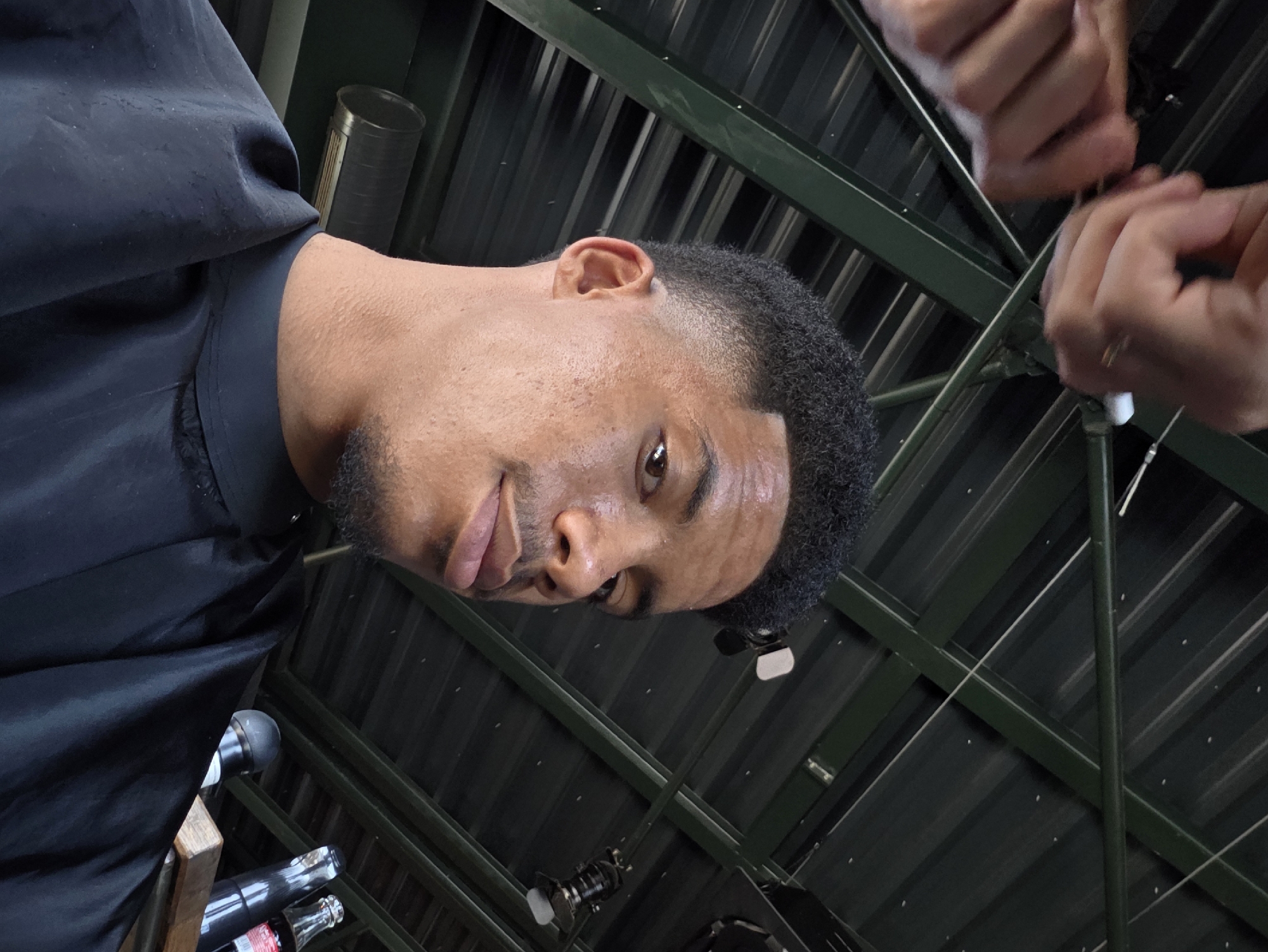 The image shows a close-up, upward-facing shot of a young Black man's face and shoulders. He is wearing a black cape, typical for a haircut. His hair is short and styled with a fade. His skin appears slightly sweaty or dewy. In the upper portion of the image, two hands are visible, one holding a tool, suggesting he is either getting a haircut or is a barber. The background consists of a dark, corrugated metal roof with visible green structural beams, suggesting an outdoor or covered outdoor setting, possibly a patio or open-air establishment. The lighting suggests it is daytime. Based on the presence of the man, the haircutting context, and the roof structure, this scene could be at an outdoor barber shop or salon in Varna, Bulgaria.