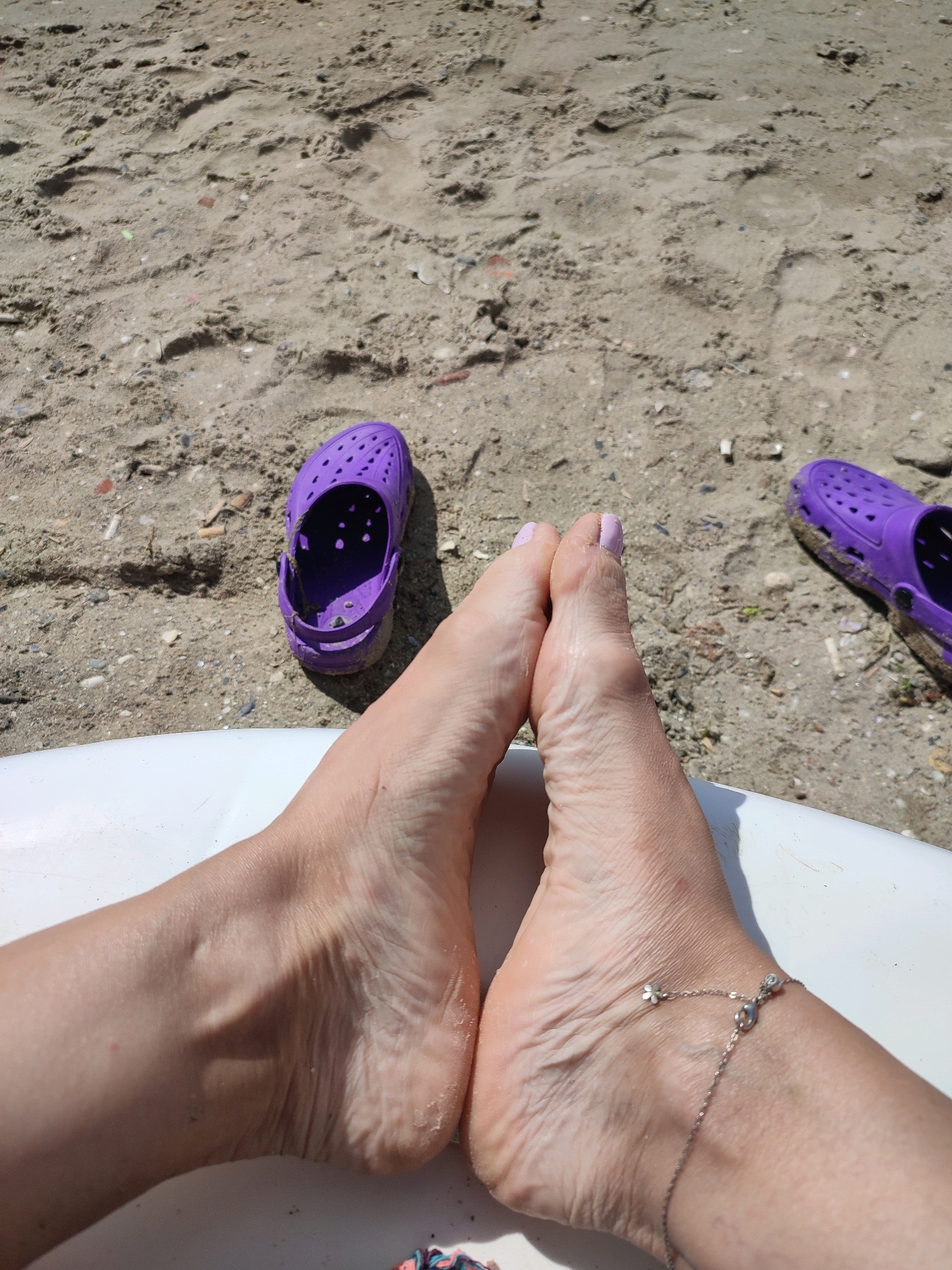 The image shows two human feet with painted toenails resting on a white surface. One foot has a silver anklet with small charms. Scattered on the sandy ground are two purple clogs, one appearing to be partially filled with sand. The background consists of a sandy area with visible footprints and small debris. The overall scene suggests a casual outdoor setting, possibly a beach. There is no information to indicate the location is Kadıköy, Turkey.