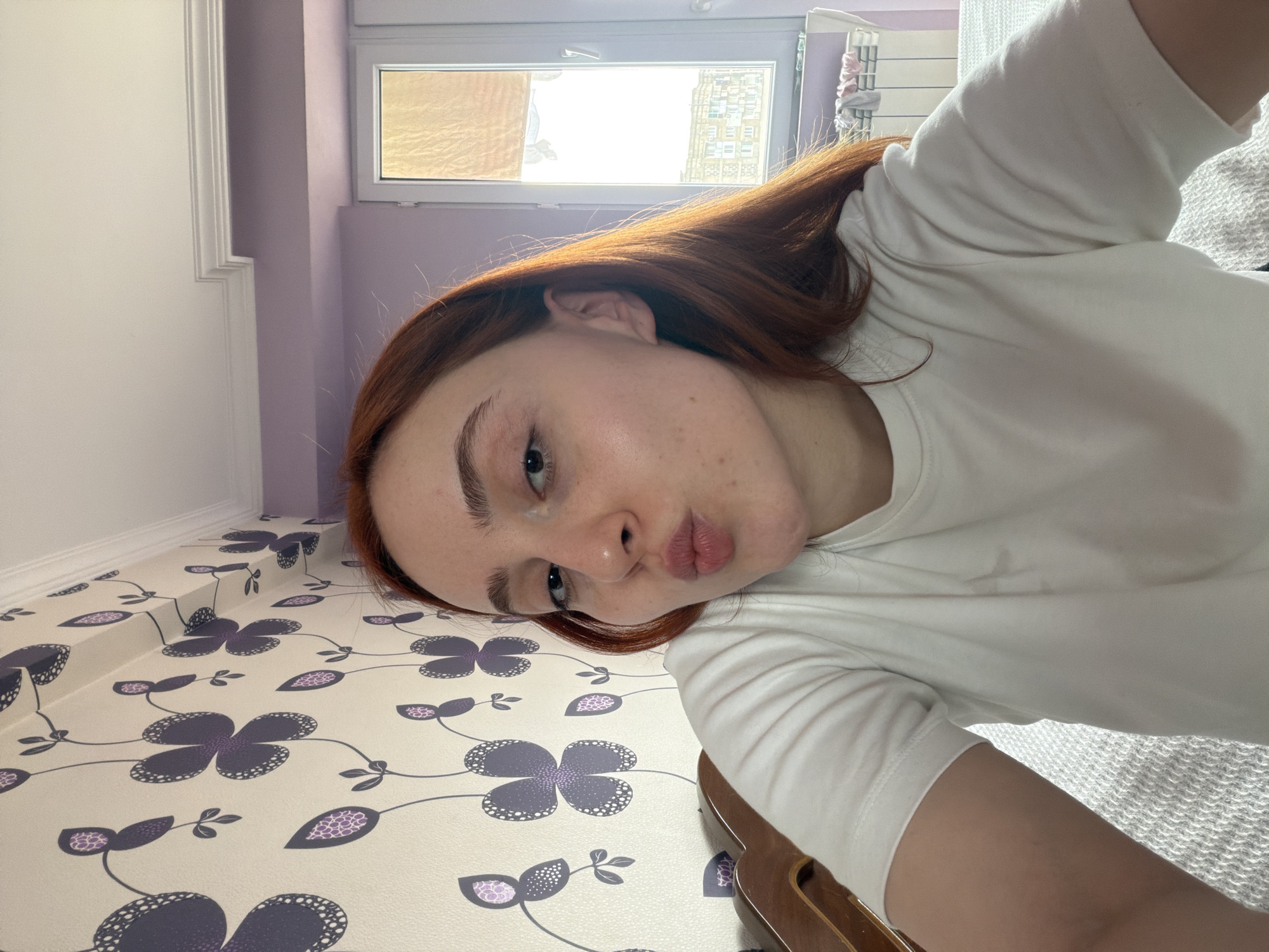 A fair-skinned person with auburn hair is posing for a selfie. The person has pursed lips. The person is wearing a white shirt. The background includes a room with lilac walls, white trim, a window, and patterned wallpaper. The location appears to be indoors, in a residential setting, but there is no visible evidence to confirm that the location is in Tbilisi, Georgia.