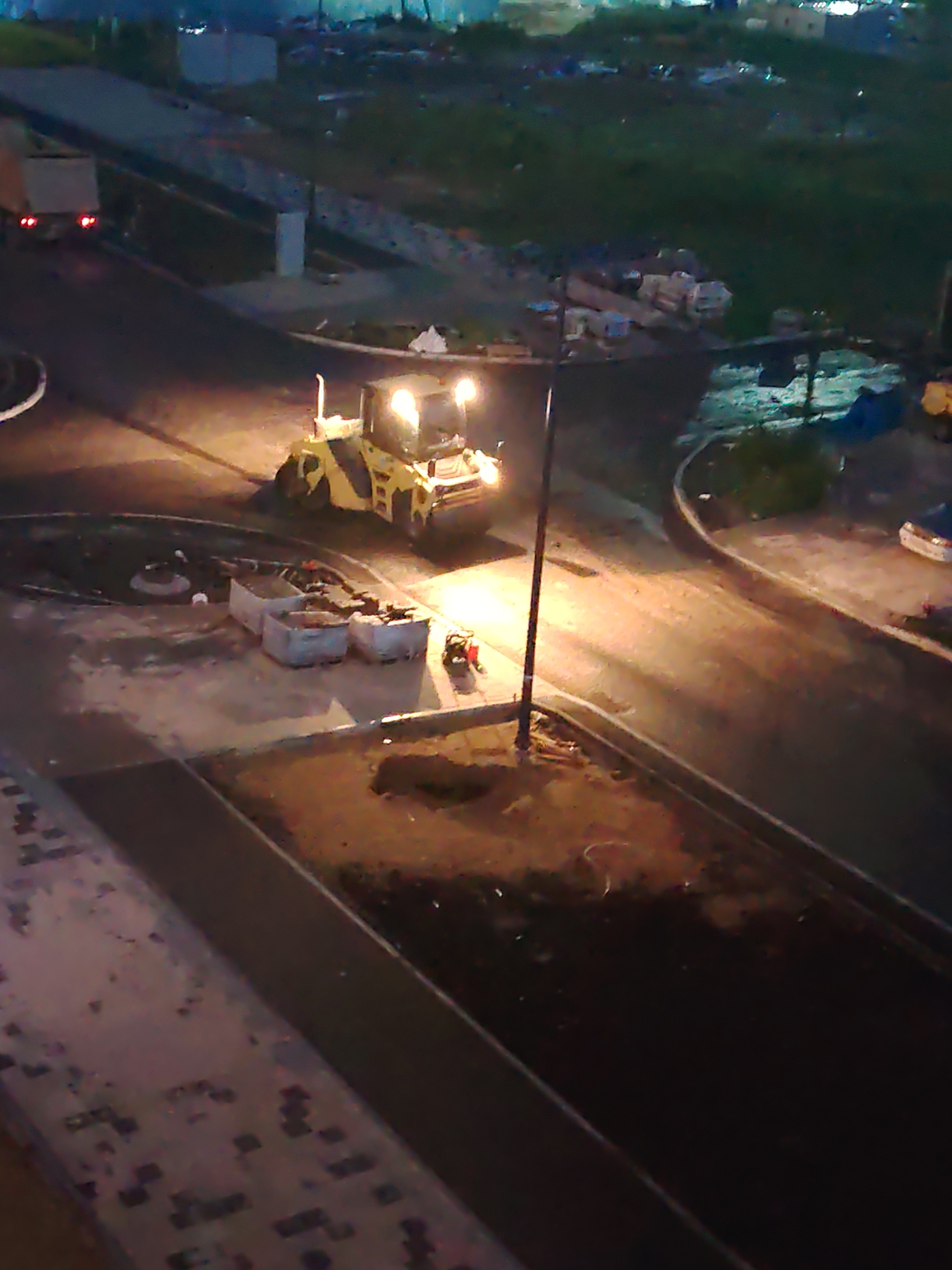 An aerial view captures a nighttime construction scene in Yekaterinburg, Russia, likely on or near a road or infrastructure project. A yellow road roller is illuminated by its own headlights and a nearby streetlamp, actively compacting asphalt. In the background, a white truck with illuminated taillights is parked. Several gray industrial bins are positioned near the road, and piles of dirt and construction debris are scattered around the area. The surrounding environment consists of dark, unlit areas with faint outlines of buildings and vegetation, suggesting an urban fringe or a developing area. The overall scene depicts ongoing road construction work under artificial lighting.