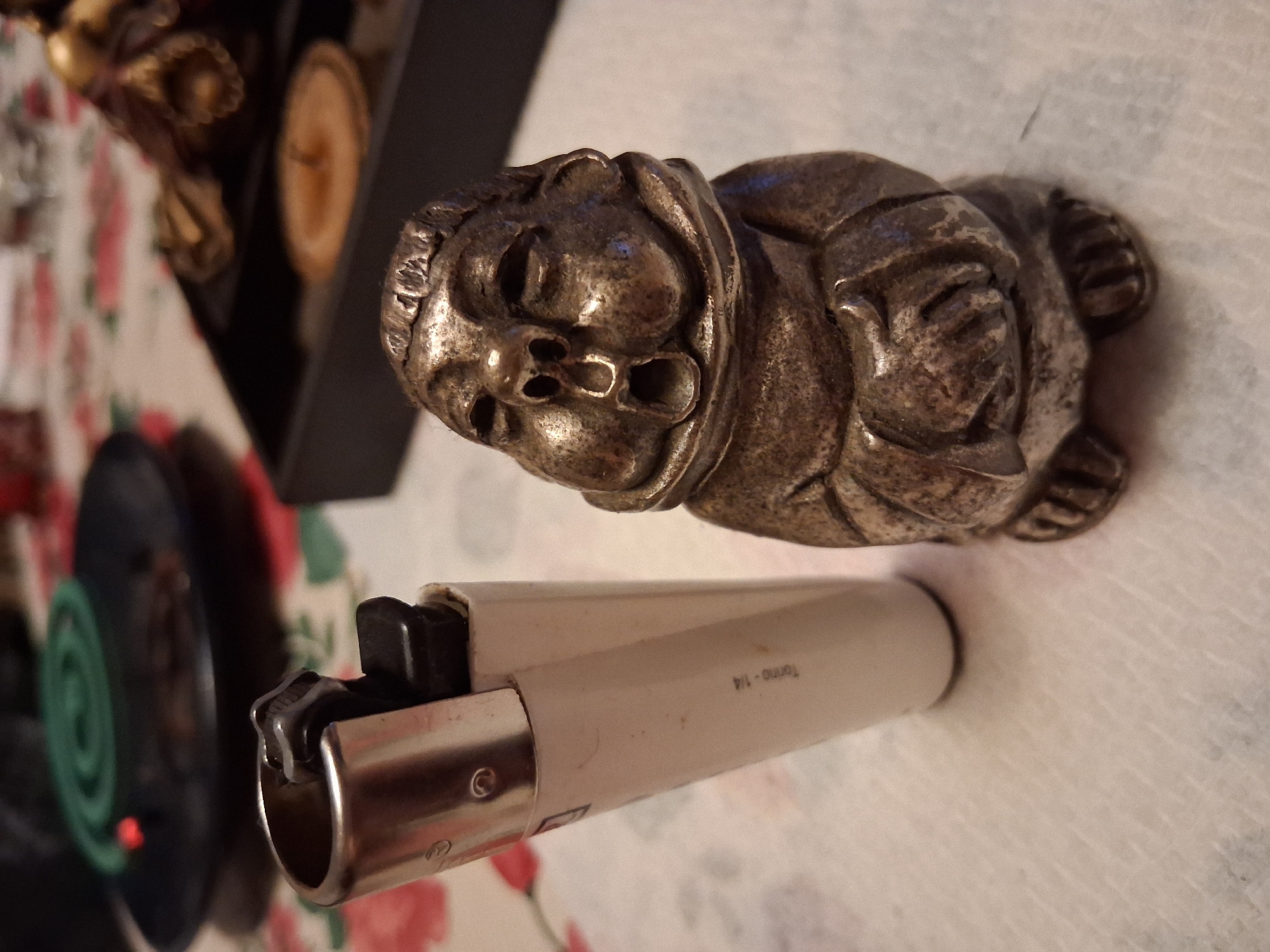A close-up shot shows a metal figurine of a gargoyle lying on a patterned tablecloth. The figurine is detailed with a grotesque face, emphasizing its features like eyes, nose, and mouth. Behind the figurine, a black rectangular object is visible, possibly a box, with two round, light-colored objects inside, resembling coasters or small decorative plates.
In the foreground, a white and silver disposable lighter rests on the tablecloth. The lighter's metallic base is clearly visible, and the white plastic body has some text imprinted on it. The text on the lighter reads "Torino - 1/4". To the left of the lighter, a dark circular object with a green spiral pattern is partially visible, with a small red light emitting from its center. This object might be an electric mosquito repellent device or a similar item.
The lighting in the image suggests it was taken indoors, possibly in the evening, as there is no indication of natural daylight. The overall scene appears to be a still life arrangement of decorative items. The setting, with the patterned tablecloth and the objects, gives a sense of a domestic or personal space. The figurine's expression could be interpreted as menacing or humorous, depending on the viewer's perspective. No specific activity is taking place; the objects are simply arranged.