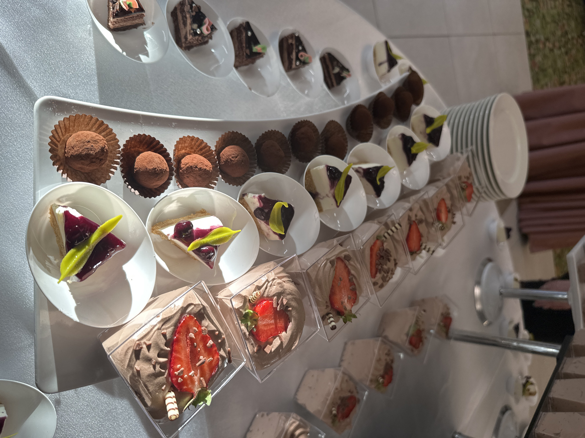 The image depicts a dessert display, likely at an event in Obrochishte, Bulgaria. The display is organized on a multi-tiered white serving stand. There are several types of miniature desserts including chocolate mousse with strawberries in small plastic cubes, small white cakes topped with dark jelly, chocolate truffles in paper liners, and slices of multi-layered chocolate cake arranged in white serving spoons. A stack of white plates is also visible on the serving stand. The image is taken at an angle, providing a top-down view of the dessert selection.