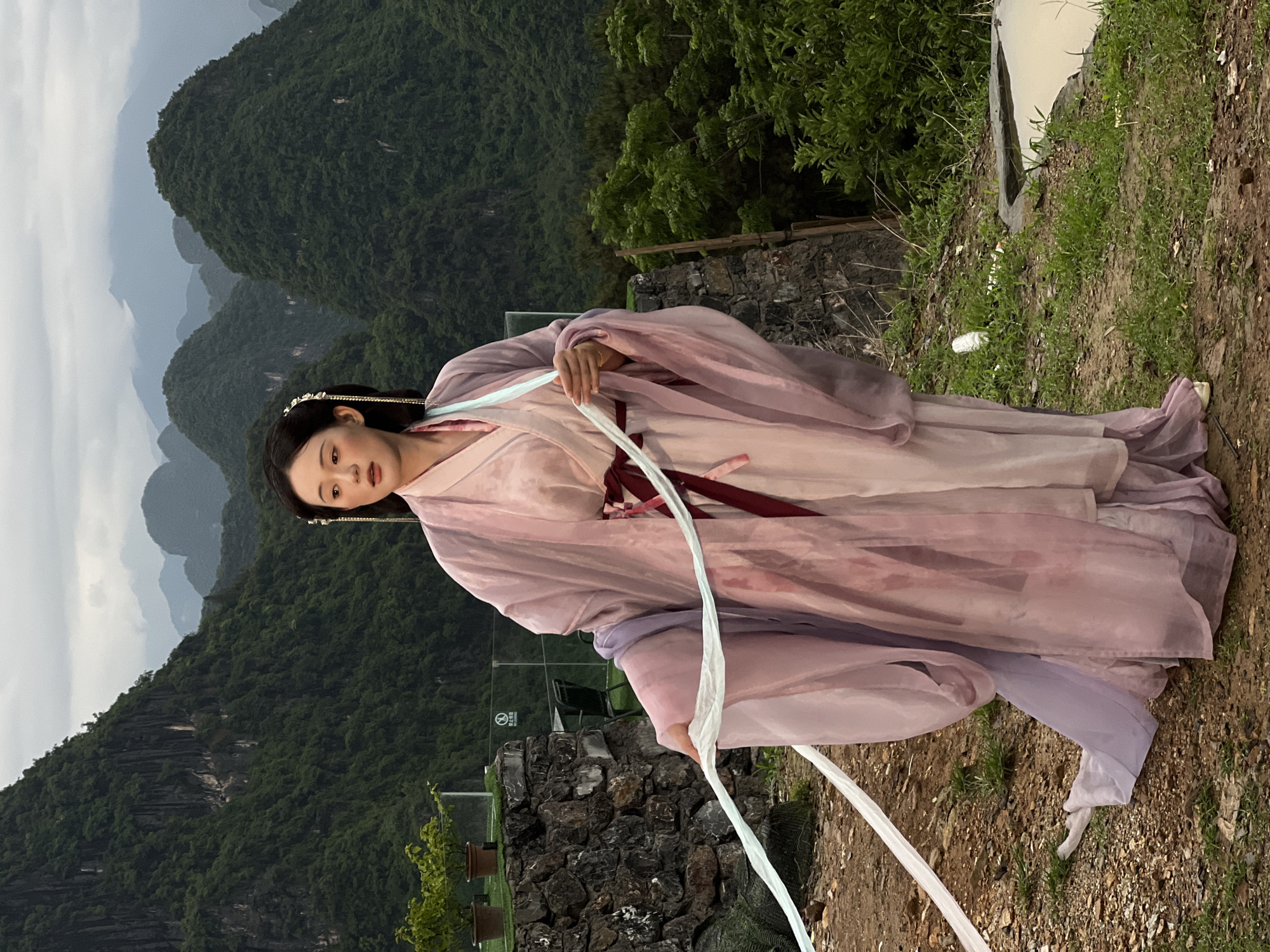 A young woman in traditional Chinese dress poses on a natural hilltop overlook in Guilin, China. She wears a pale pink and purple Hanfu-style robe with long, flowing sleeves and a maroon sash, paired with a white fabric streamer she holds loosely. The woman has dark hair adorned with a decorative hairpiece. The background features the iconic karst mountains of Guilin, partially obscured by haze. The foreground consists of a rough, earthen terrain and a low stone wall. The overall scene creates a sense of historical charm and harmony with the natural landscape.