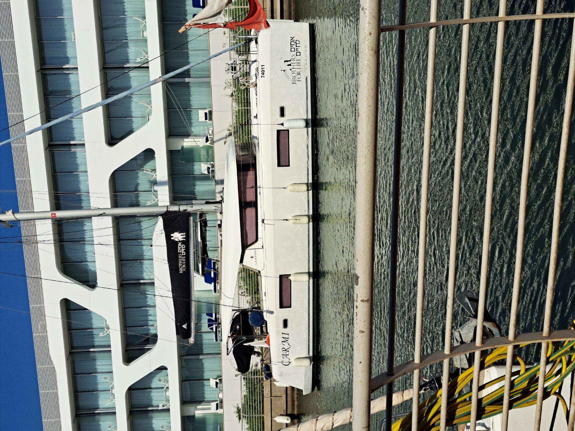 The image shows a white catamaran sailboat docked in a marina in Herzliya, Israel. The boat is named "CARMI" and displays the words "BROTHERS FOR LIFE" in both Hebrew and English, along with a silhouette of two people. Behind the boat is a modern white building with a distinctive curved facade and many windows. The sky is clear and blue. In the foreground, a metal railing with horizontal bars obscures part of the view of the water and the boat's hull. The water is dark green and reflects the sunlight, showing ripples. Various nautical equipment, such as ropes and fenders, are visible on and around the boat.