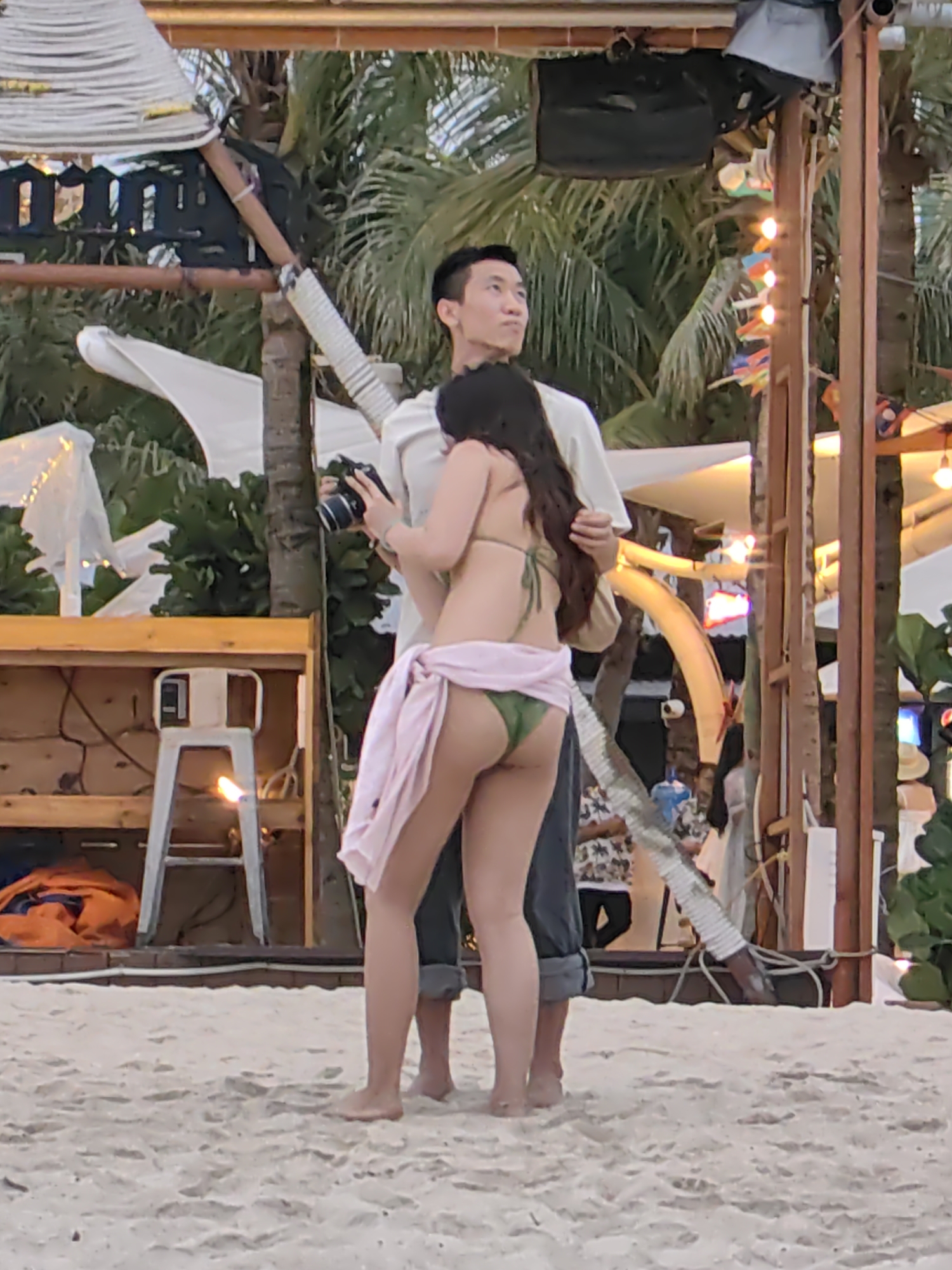 This image captures a beach scene in Bai Chay Ward, Vietnam, likely during the evening or late afternoon, given the warm ambient lighting and illuminated string lights. The primary subjects are a man and a woman standing barefoot on the sand.
The woman is positioned in the foreground, facing away from the camera, revealing her long dark hair. She wears a green bikini bottom and a light pink sarong or wrap around her hips. She is holding a black DSLR-style camera in her hands. Behind her, slightly to the left, stands a man with short dark hair, wearing a light-colored long-sleeved shirt and dark cuffed pants. He is looking towards the right side of the frame.
The background features typical beach elements, including numerous green palm trees. There are also wooden and bamboo structures, draped with white fabric and decorated with string lights, suggesting a festive or recreational area such as a beach bar or restaurant. To the left, a wooden counter or table is visible, in front of which stands a white stool. On the stool, there is a prominent glowing light source, possibly a torch or a small fire, contributing to the evening atmosphere. An orange bundle of fabric is on the ground near the counter. The sand appears clean and well-trodden. No discernible text is visible within the frame.