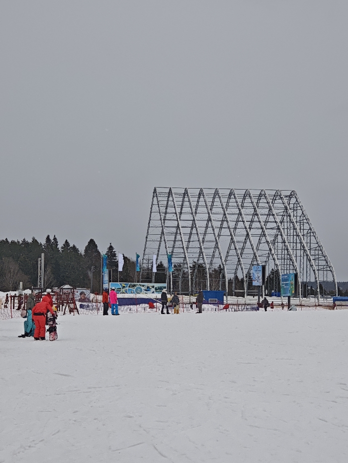 The image displays an outdoor winter scene at a snow-covered facility in Курово, Russia, under a uniformly overcast, light gray sky. In the foreground, left of center, a person wearing a red snowsuit stands next to a snowboard lying flat on the snow. Another individual, dressed in a teal jacket and dark pants, is seated directly on the snow nearby. The mid-ground features multiple groups of individuals in winter attire, standing on the snow. Visible objects in this section include red barriers, a small wooden-toned structure resembling playground equipment, and several tall banners and flags, some white and others blue with graphics, positioned in front of a dense pine forest line. To the right and extending into the background, a large, gray metallic truss structure, composed of multiple interconnected A-frame segments, dominates the horizon. A single blue container or temporary building is discernible beneath this structure in the mid-ground. The entire ground surface is covered in white snow, exhibiting some minor surface indentations.