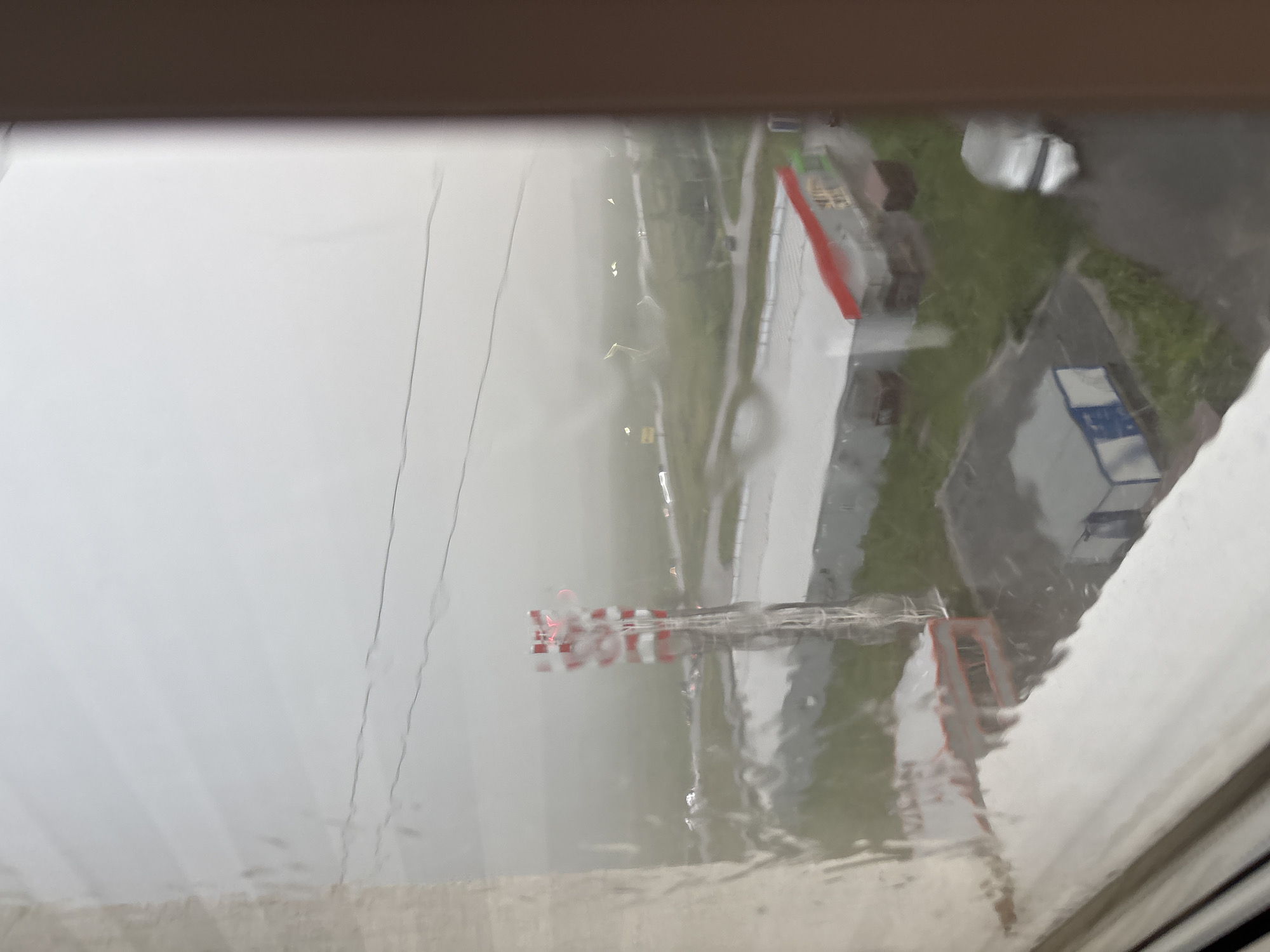 The image is a blurry, overcast outdoor scene viewed through a wet window. In the middle ground, a tall, red and white striped tower, possibly a communication tower, stands prominently. To the right of the tower is a long, low-rise building with a red roof and some green accents, appearing to be a commercial or industrial structure. Further to the right, a silver vehicle is parked. In the foreground, water streaks on the window obscure the view. The background consists of a hazy, indistinct landscape with what appear to be roads and distant lights. The overall impression is one of a dreary, possibly rainy day in an industrial or semi-urban area. This scene is located in Засечное, Russia.