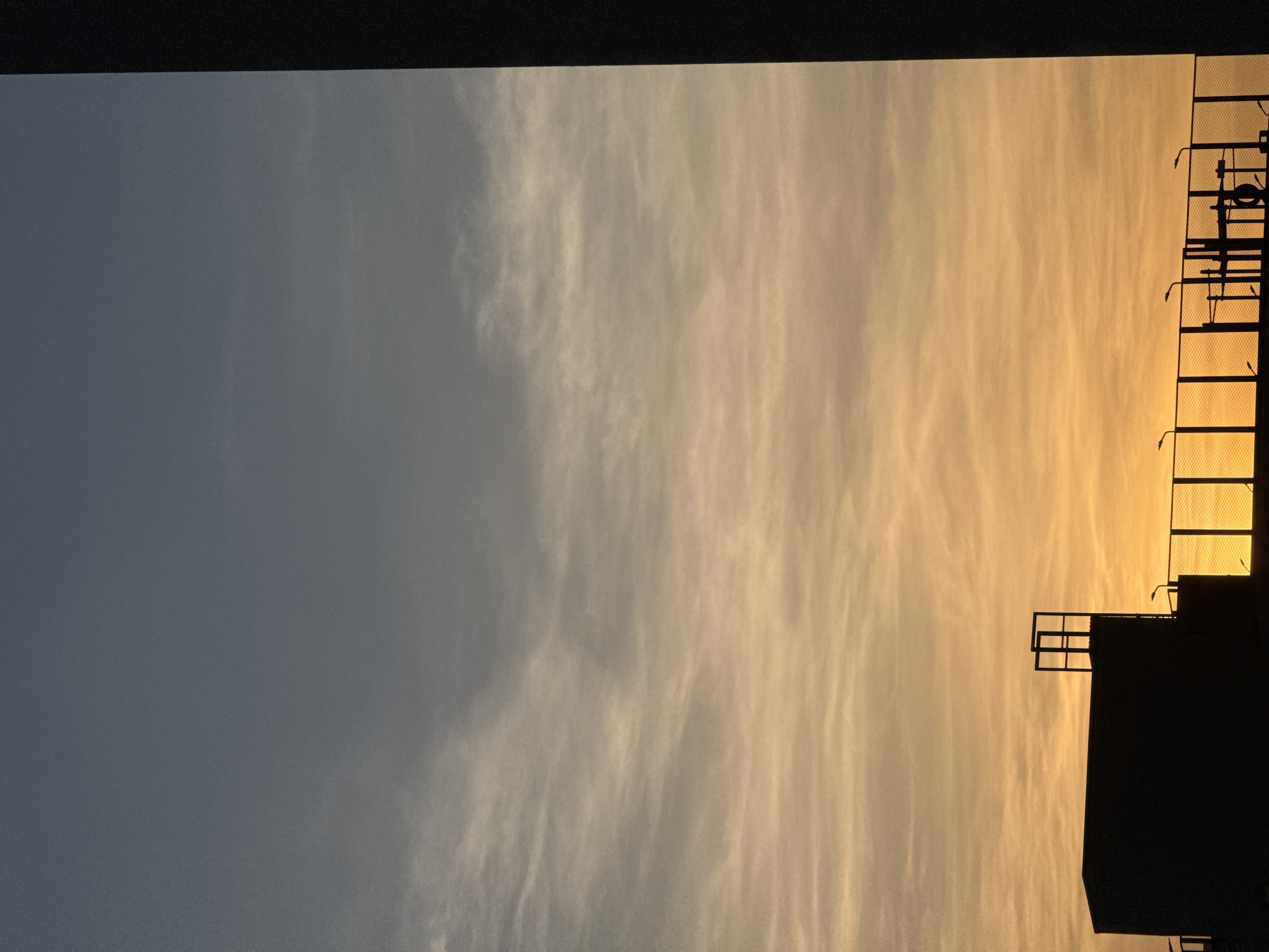 The image shows a dusk or dawn sky in Yekaterinburg, Russia. The sky transitions from a dark blue-grey to a hazy orange-yellow. Thin, wispy clouds are visible. Silhouetted against the sky is a building or structure with a ladder extending upwards. The structure has a platform with railings at the top.
