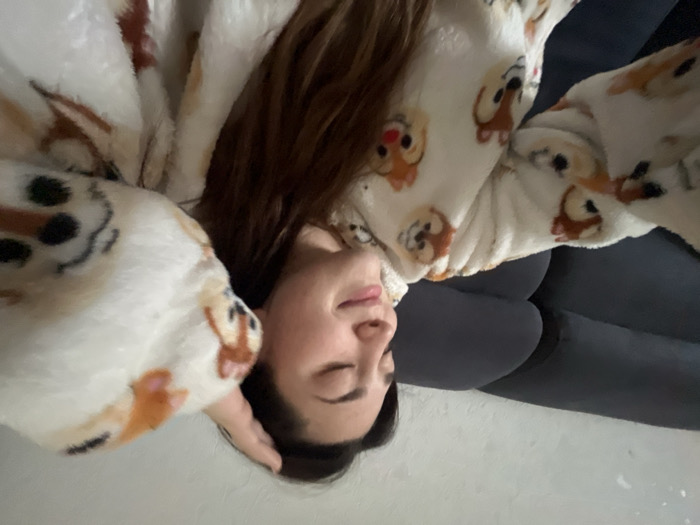 An indoor scene in a private residence in Rennes, France, depicts a young adult female. She has long, dark brown hair and is wearing a cream-colored, plush hooded garment patterned with various cartoon characters, including Chip and Dale and Olaf. Her eyes are closed, and she displays a subtle smile. Her right hand is raised, with fingers touching the top of her head. A dark grey sofa is visible behind her, and a light-colored, textured wall constitutes the upper background.
