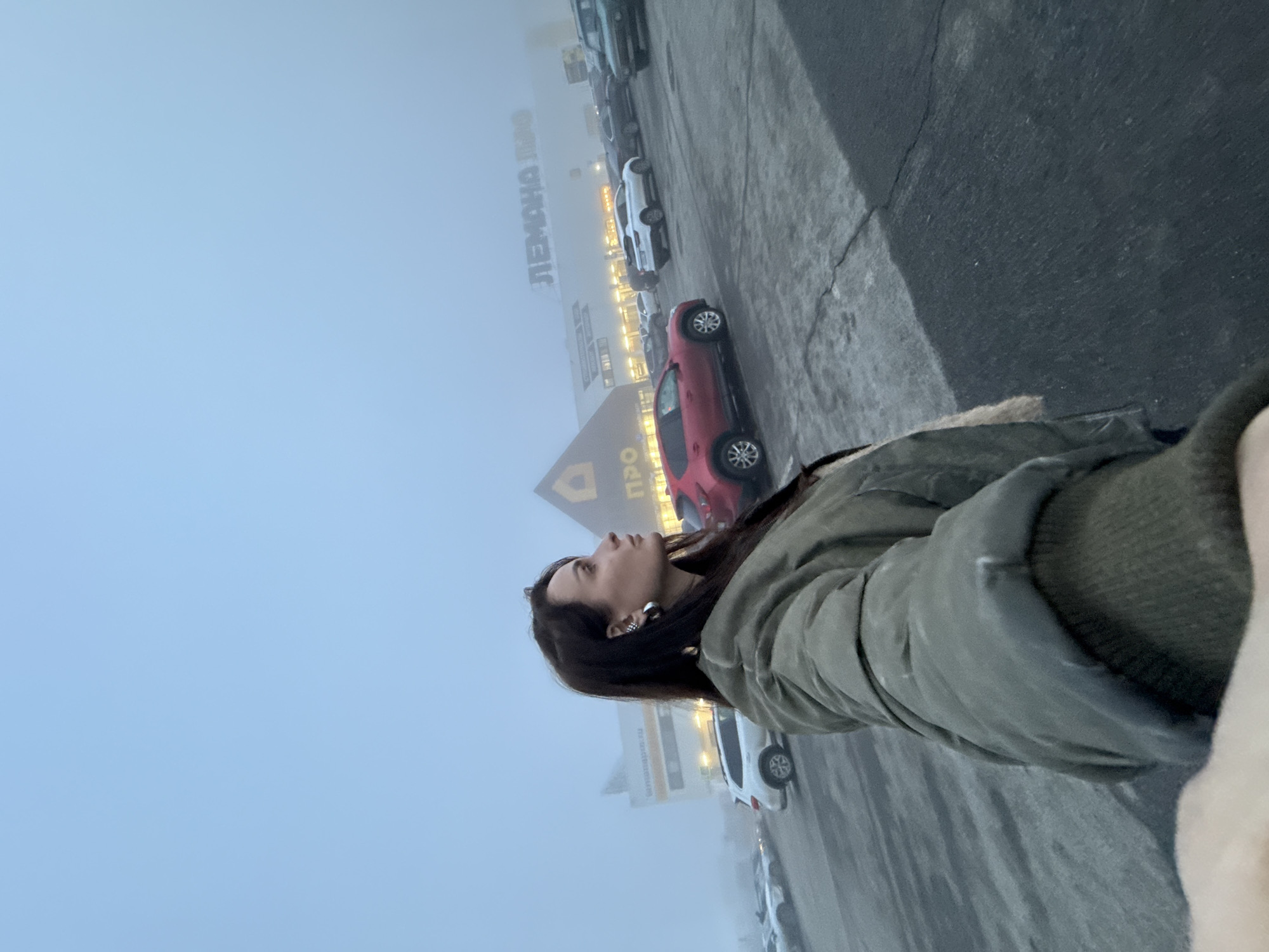 A woman stands in a parking lot, looking up at a building in a city engulfed in heavy fog. The building is identified by signage written in a Cyrillic alphabet and appears to be a commercial establishment called "LEMANA PRO". The location is in Izhevsk, Russia. The woman wears a green puffer jacket and has dark hair. Several parked cars are visible in the parking lot. The asphalt ground shows wear and tear.