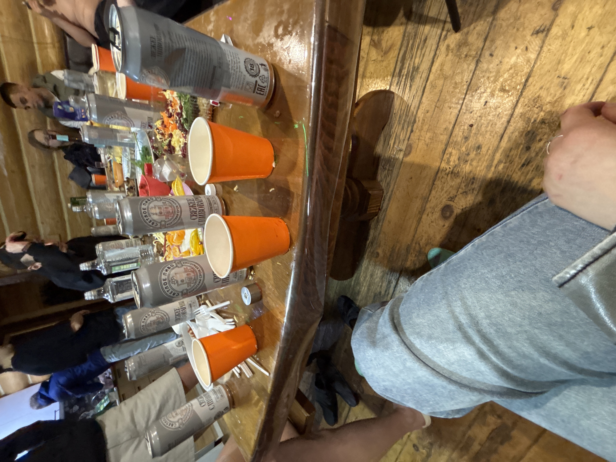 The image captures a social gathering in what appears to be a rustic setting, potentially a home or cabin, likely located in the city of Chaikovsky, Russia given the branding on the beverages. People are visible around a wooden table laden with various items, including cans of a drink labeled "СВЕЖЕЕ ПИВО СВЕТЛОЕ" (Fresh Light Beer) which suggests its Russian origin, possibly from the local brewery. There are bottles, salads, and orange plastic cups scattered on the table, indicating a casual party or meal. The focus is primarily on the table and its contents, while individuals around the table are seen from varying angles, some more distinct than others. A person wearing jeans is partially visible on the right side of the image. The overall environment suggests a relaxed, informal social event.