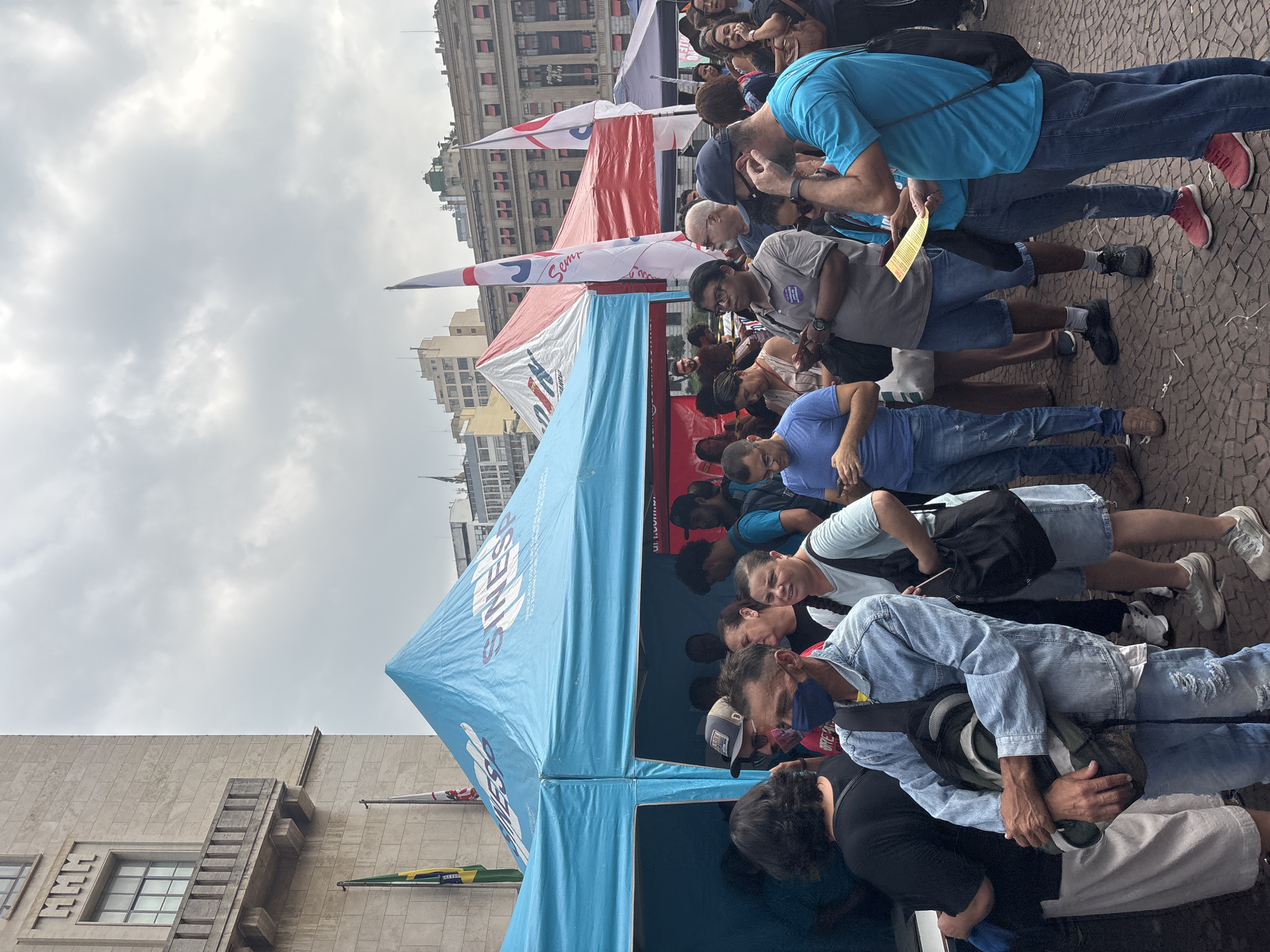 This image depicts an outdoor public gathering in São Paulo, Brazil, under an overcast sky during the daytime. The scene is set on a paved street or square, with several multi-story city buildings forming the backdrop.
Numerous people, primarily adults, are visible, standing and engaging in various activities. They are casually dressed in t-shirts, jeans, and shorts. Some individuals appear to be looking at papers or phones, while others are conversing. A man in a blue shirt is holding a yellow piece of paper. The overall mood appears attentive and engaged, suggesting participation in an organized event.
In the foreground and midground, two large tents are set up: a prominent blue tent and a red tent behind it. The blue tent features the prominent text "SINESP" along with partial visibility of "SIND..." and "SINDICATO," suggesting an affiliation with a union or syndicate. Its logo appears to include a stylized depiction of figures or a handshake. Several flags are also present, including white flags with red accents that display text such as "Sempre..." and "Sem lutar..." A Brazilian flag is partially visible near the base of the blue tent.
The surrounding buildings exhibit typical urban architecture, with multiple windows and balconies. The paved ground, possibly cobblestones, is visible beneath the feet of the attendees. The combination of tents, flags, and an assembled crowd indicates a public event, possibly a rally, informational session, or community gathering.