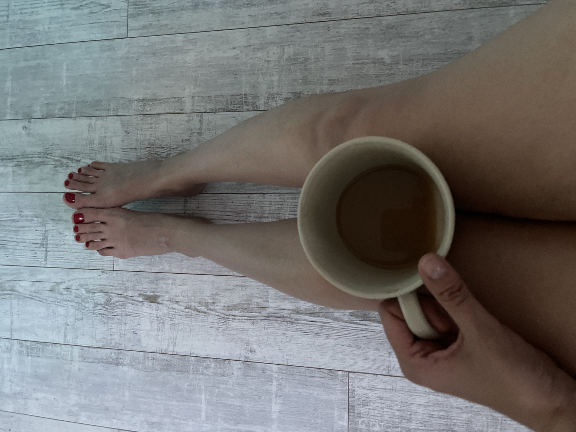A person's legs are positioned on a weathered, light-colored wooden floor. The feet, with red painted toenails, are visible on the left side of the frame. A hand is holding a beige ceramic mug with a tan liquid inside, positioned in the center of the frame against the person's legs. The scene is indoors, and no information is available regarding the location's city or environment.