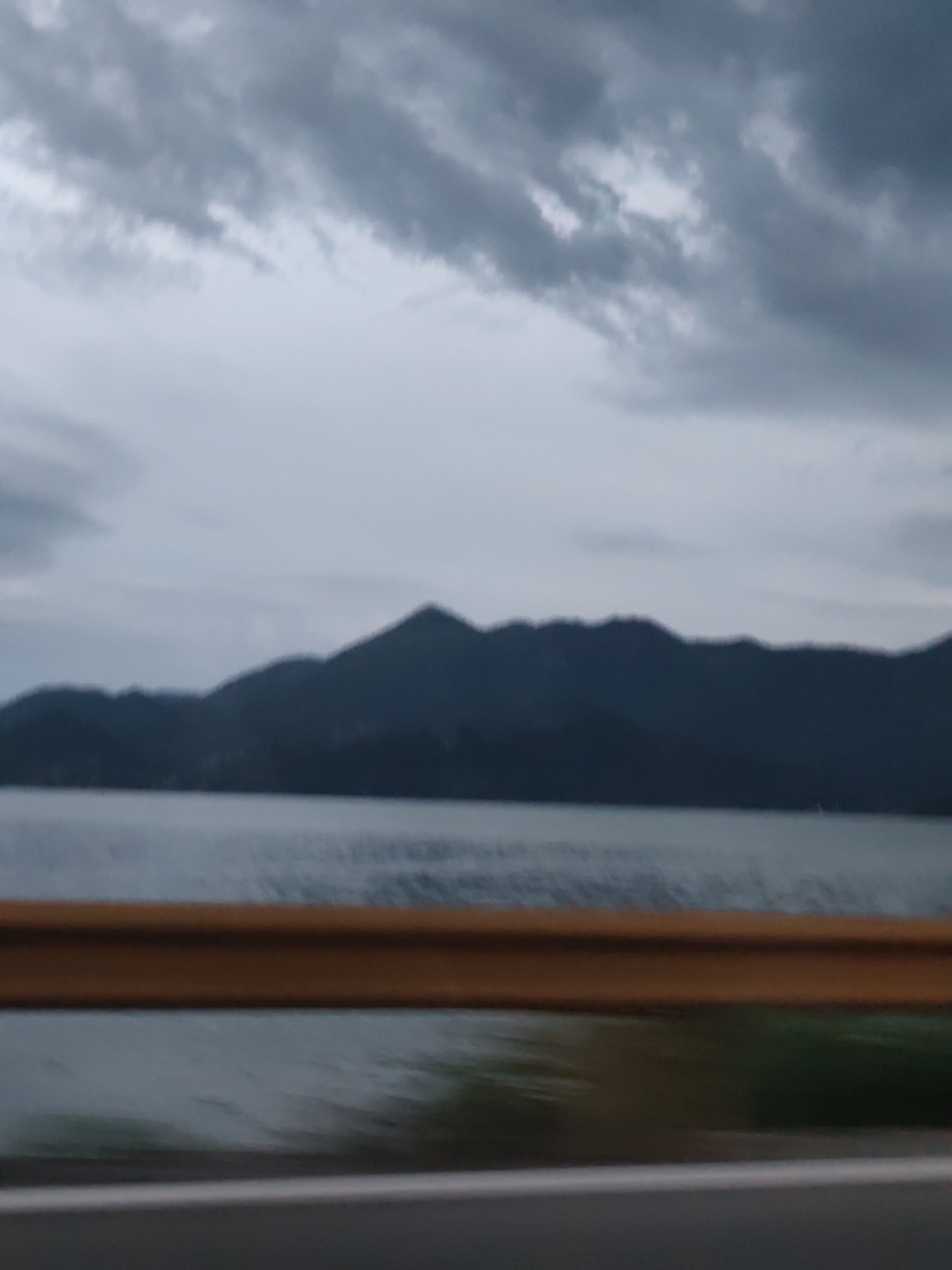 A wide shot captures a slightly blurred, overcast sky over a calm body of water with a dark, silhouetted mountain range in the distance. In the foreground, a bright orange guardrail is visible across the bottom third of the image, with a grey road surface below it. The scene appears to be located near Vranjina, Montenegro, as suggested by the mountainous landscape and body of water, likely Skadar Lake. No people or specific objects are discernible due to the blurriness and distance. The overall mood is somber, likely due to the cloudy weather.