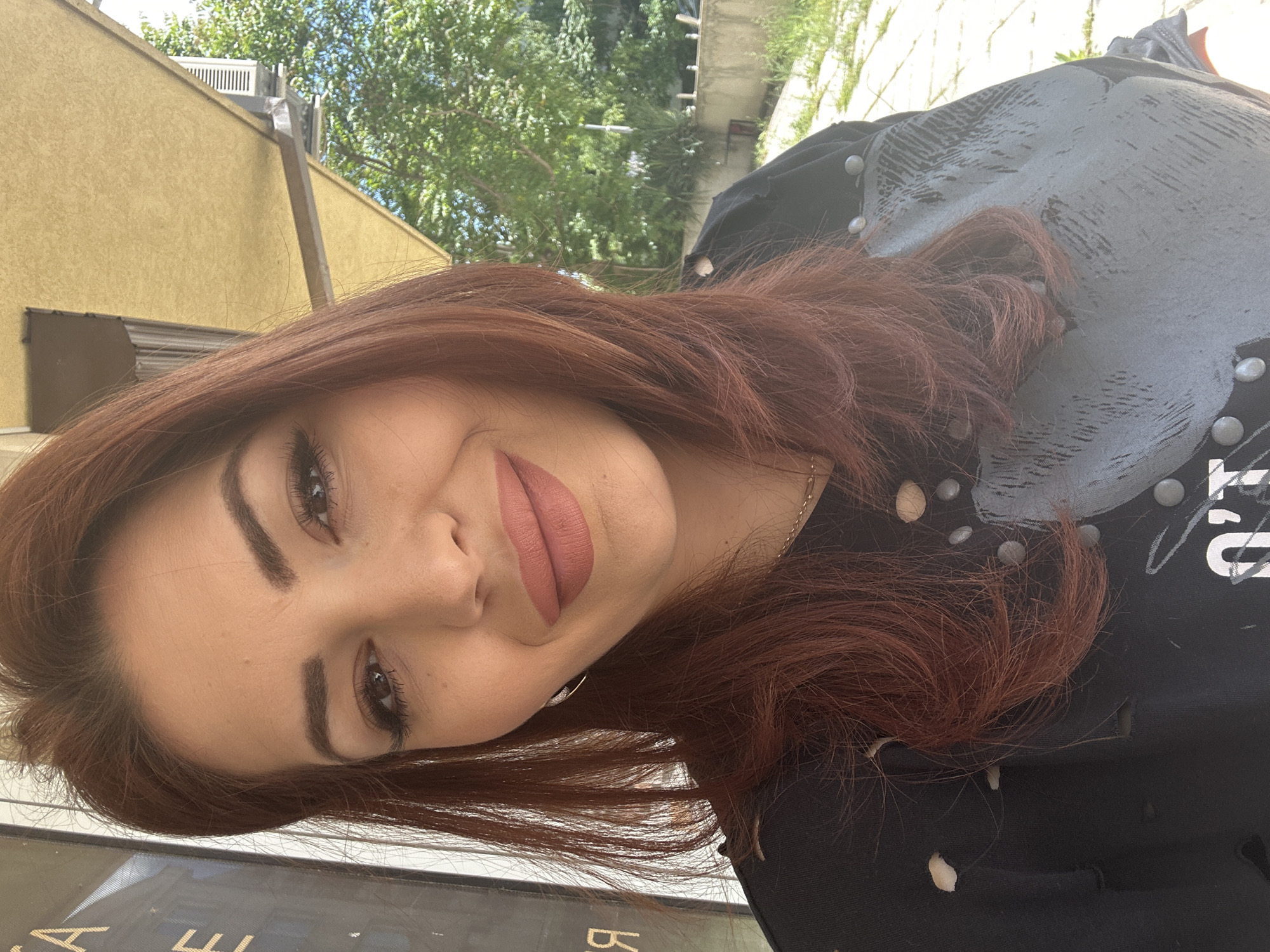 A person with auburn hair is featured in a close-up selfie. They have fair skin, dark eyebrows, and brown eyes. They are wearing lipstick with a matte finish in a neutral tone. The person is wearing a black shirt with a gray design, adorned with silver studs.
The background includes a beige building and green trees. The photo appears to be taken outdoors in a city environment, but the image does not contain enough information to ascertain that the location is Sofia, Bulgaria.