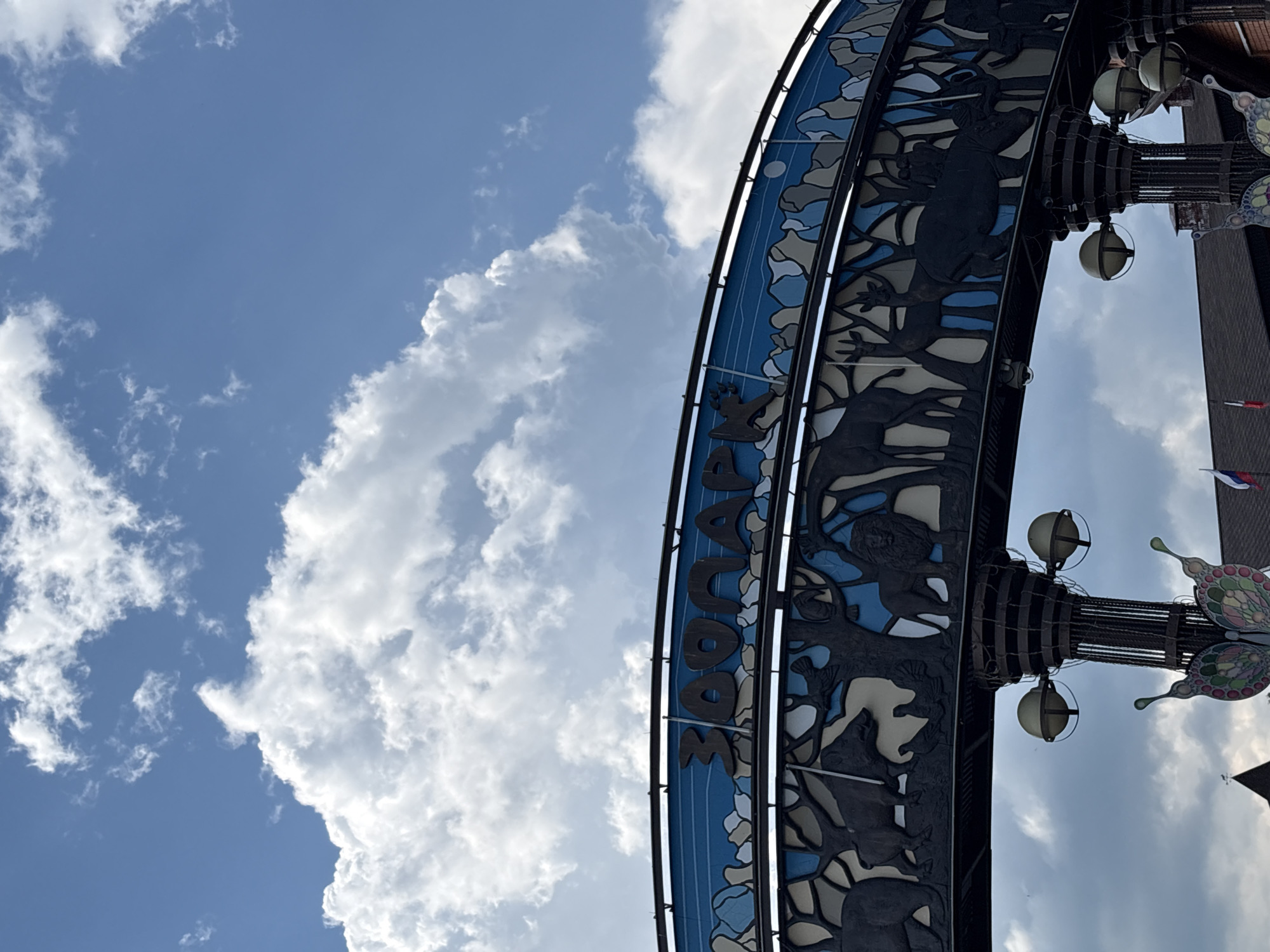The image shows an arched structure with the word "ЗОOПАРК" (ZooPark) prominently displayed in large, dark letters against a blue background with white and grey rock-like shapes. Below the word, a silhouetted frieze depicts various animals, including a bear, boar, birds, a lion, deer, and what appears to be a rhinoceros or elephant. The arch is supported by dark, cylindrical structures with lamps on them. In the foreground, below the supports, are decorative elements resembling butterflies. In the background, there is a cloudy blue sky. The architecture and the Russian text indicate the location is likely the Izhevsk Zoo in Izhevsk, Russia.