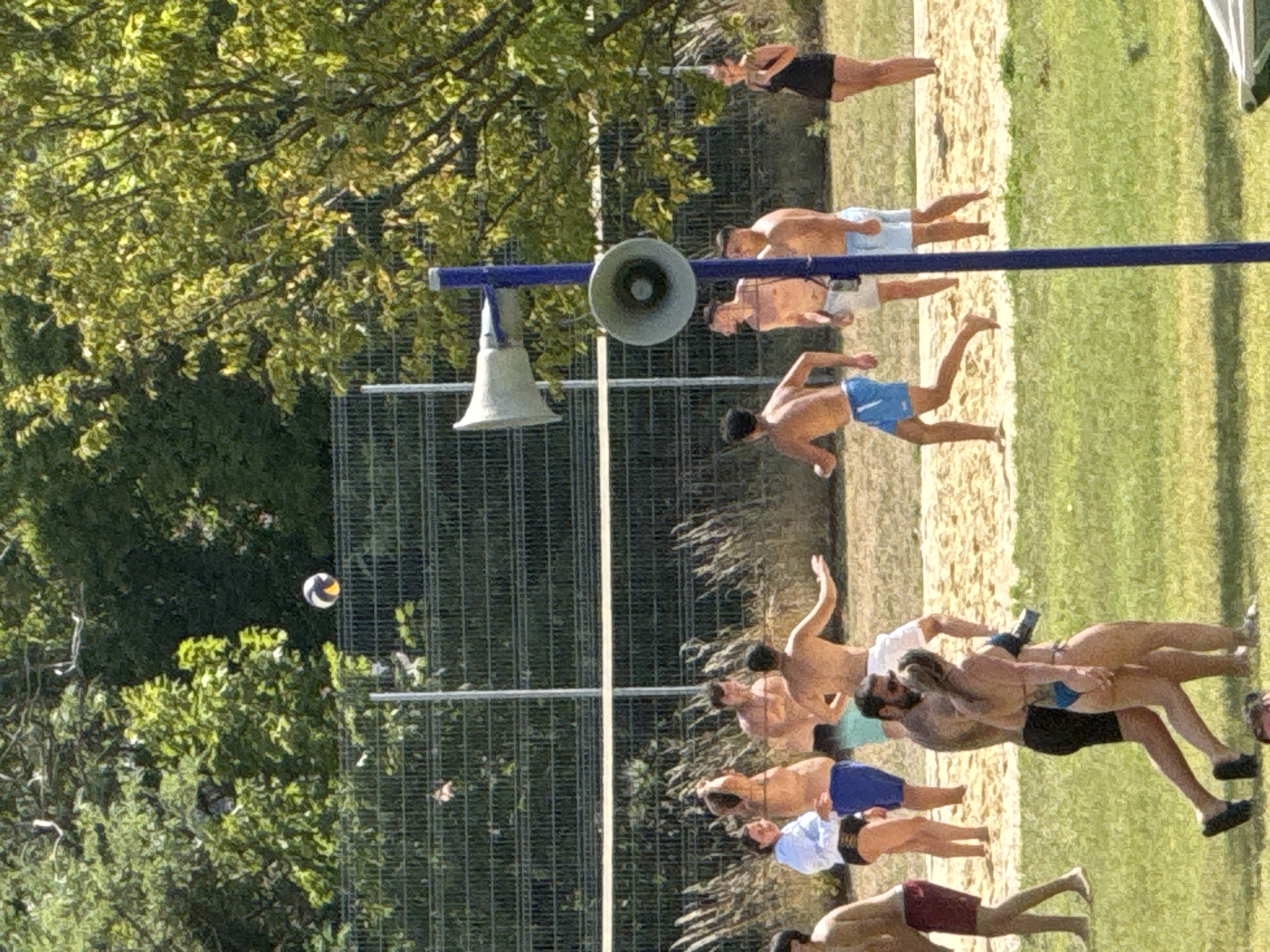 The image depicts a group of people playing volleyball on a sandy court, with a net stretched across the middle of the court. The scene is set outdoors, with lush green trees and vegetation in the background, suggesting a park or recreational area. The sky is bright, indicating a sunny day. A blue pole with two speakers is prominently positioned in the foreground, to the right of the volleyball court. The people are casually dressed in swimwear, with some actively engaged in playing the game and others observing. The overall atmosphere appears to be relaxed and recreational. This scene is likely taking place at a beach volleyball court located in Hainburg an der Donau, Austria, known for its recreational facilities.