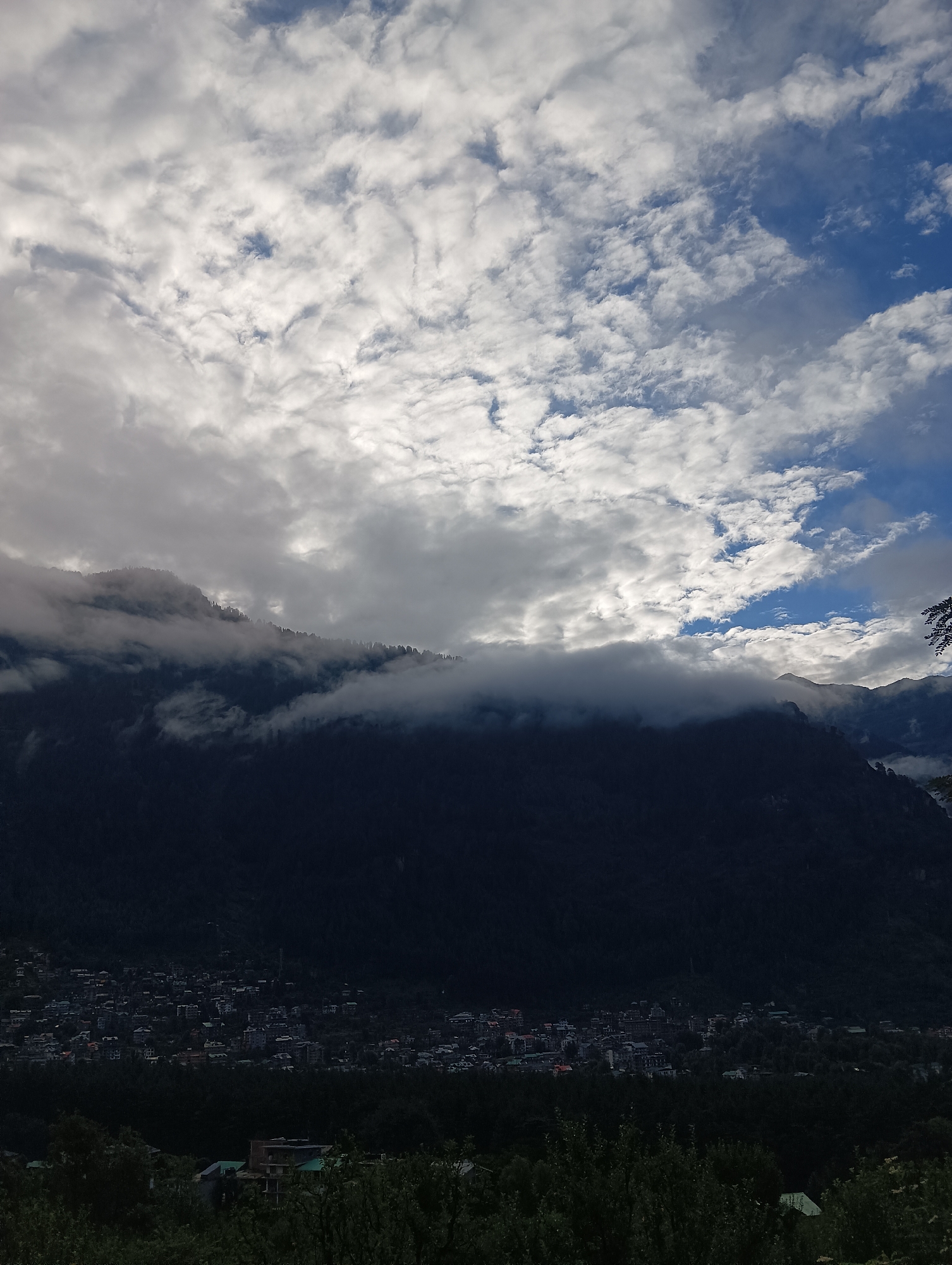 The image depicts a panoramic view of the city of Manali, India, nestled within a valley of densely forested mountains. The sky is a dramatic canvas of white clouds, with patches of brilliant blue peeking through. These clouds appear to be partially obscuring the mountain peaks, giving them a mystical, ethereal quality. In the foreground, lush green trees frame the lower portion of the image, suggesting an elevated vantage point from which the photograph was taken.
The mid-ground is dominated by the dark silhouette of the mountains, their slopes covered in dense vegetation. A town is visible in the valley below, its buildings scattered amongst the trees. The architecture suggests a mix of traditional and modern construction, with some structures featuring red or green roofs. The town appears to be spread out along the base of the mountains, illuminated by the soft light of the sky. The overall impression is one of a serene and picturesque landscape, with the natural beauty of the mountains and sky contrasting with the man-made structures of the town.
