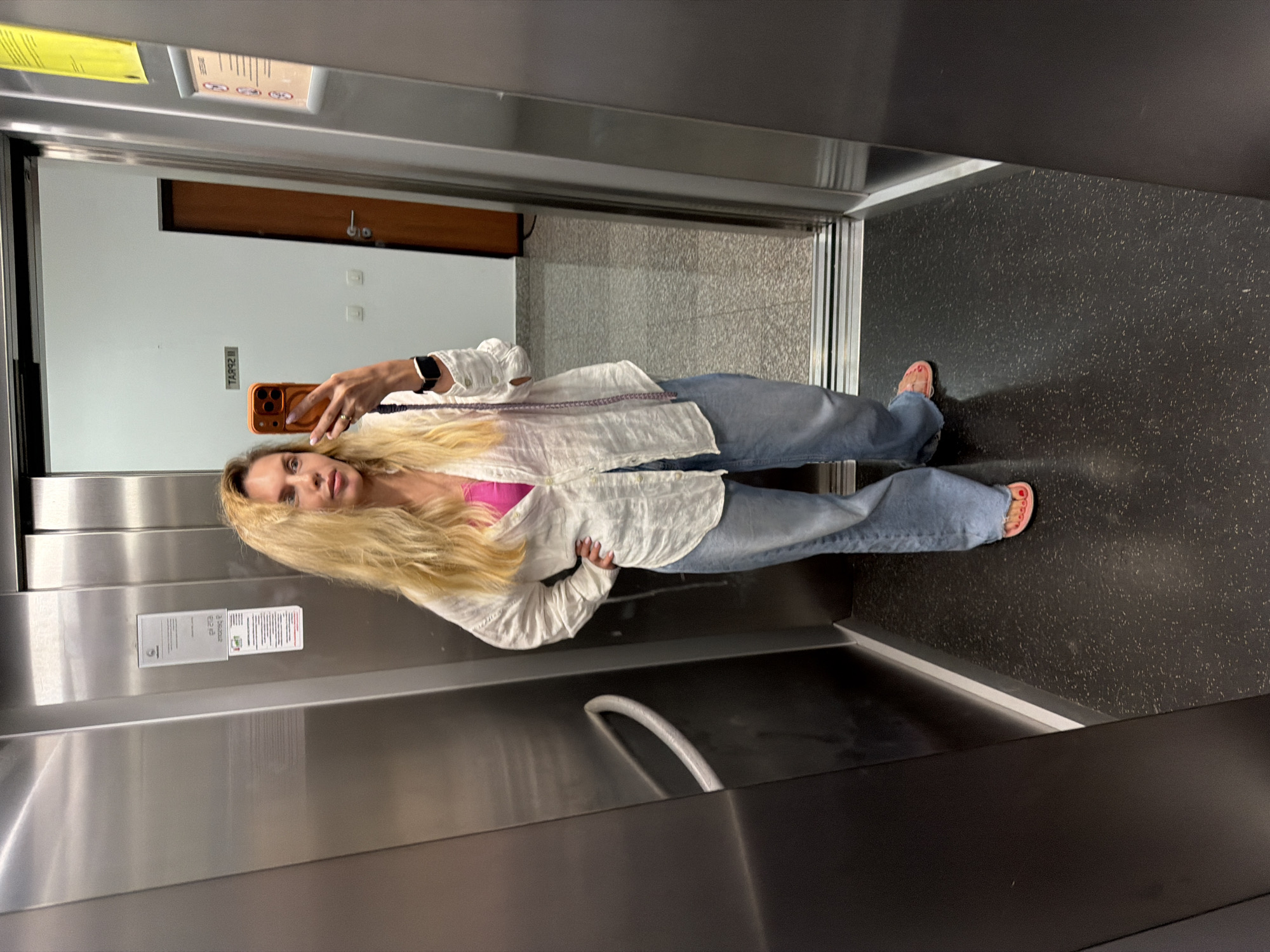 A woman with long blonde hair is taking a mirror selfie inside what appears to be an elevator in Budva, Montenegro. She is standing upright, holding an orange smartphone with multiple cameras (likely an iPhone) with her left hand, which also bears a watch and a ring. She wears a white long-sleeved shirt unbuttoned over a bright pink top, light blue jeans, and pink flip-flops.
The elevator interior features brushed metal panels on the walls and a dark speckled floor. A white curved handrail is visible on the lower right side of the frame. Reflected in the mirror, behind the woman, is a hallway with a wooden door, a white wall, and possibly light switches. Several signs or notices are affixed to the elevator wall, reflected in the image. One sign, mirrored, shows partially legible text that could include "2 ZAKONA". Another sign has smaller, illegible text and some pictograms. A yellow sticky note with illegible text is also reflected on the upper left. The woman's expression is neutral as she looks towards her phone. The setting suggests an indoor environment, likely a building's common area.