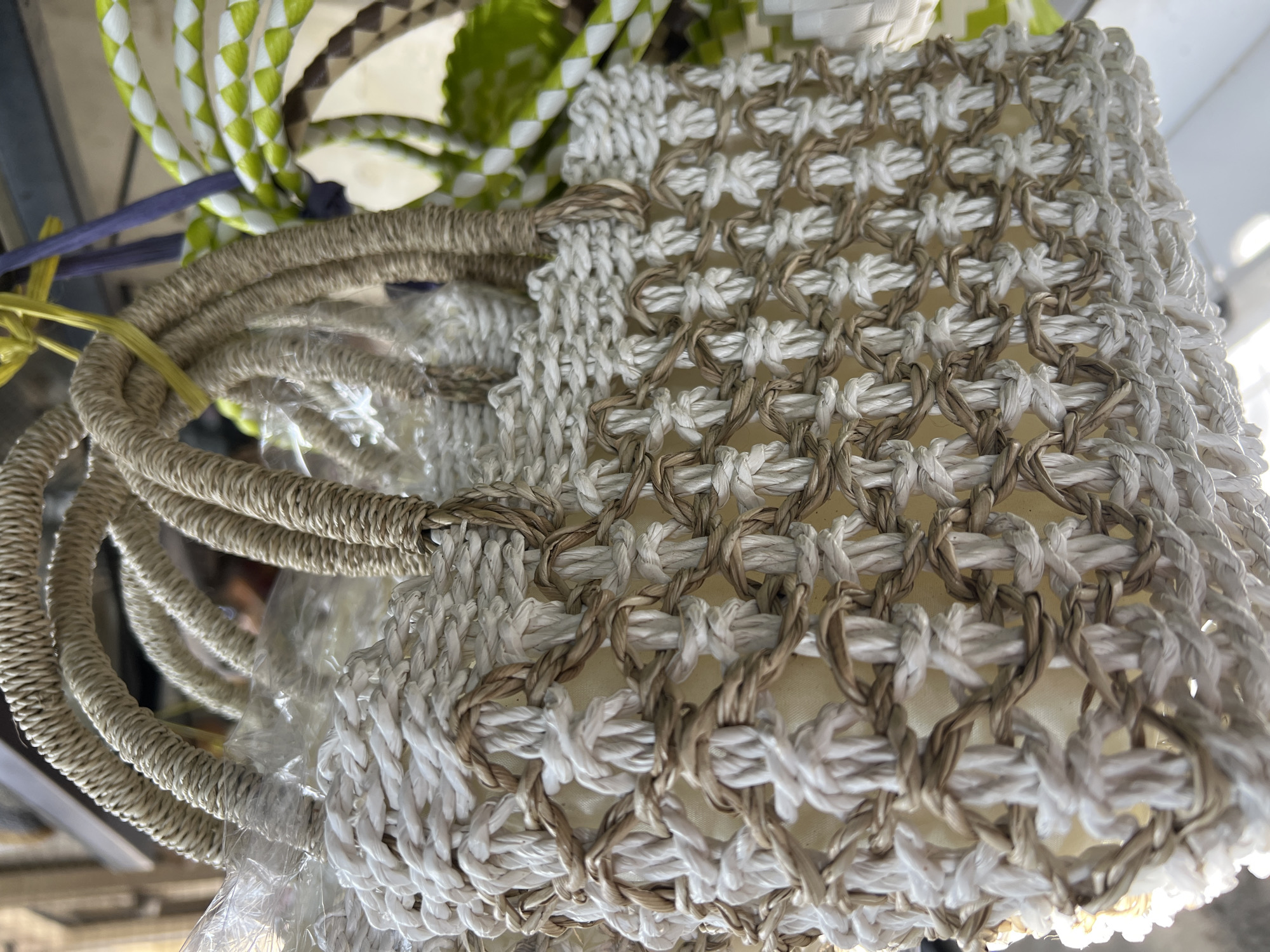 The image shows a close-up of a handmade macrame tote bag in Singakerta, Indonesia. The bag features a woven pattern of white and brown rope, forming a diamond-shaped grid. The bag has thick rope handles, likely made of the same brown material. Green and white striped rope is visible in the background. The bag has an off-white liner inside of it. Plastic wrap is partially covering the handles. The tote bag is in an indoor setting.