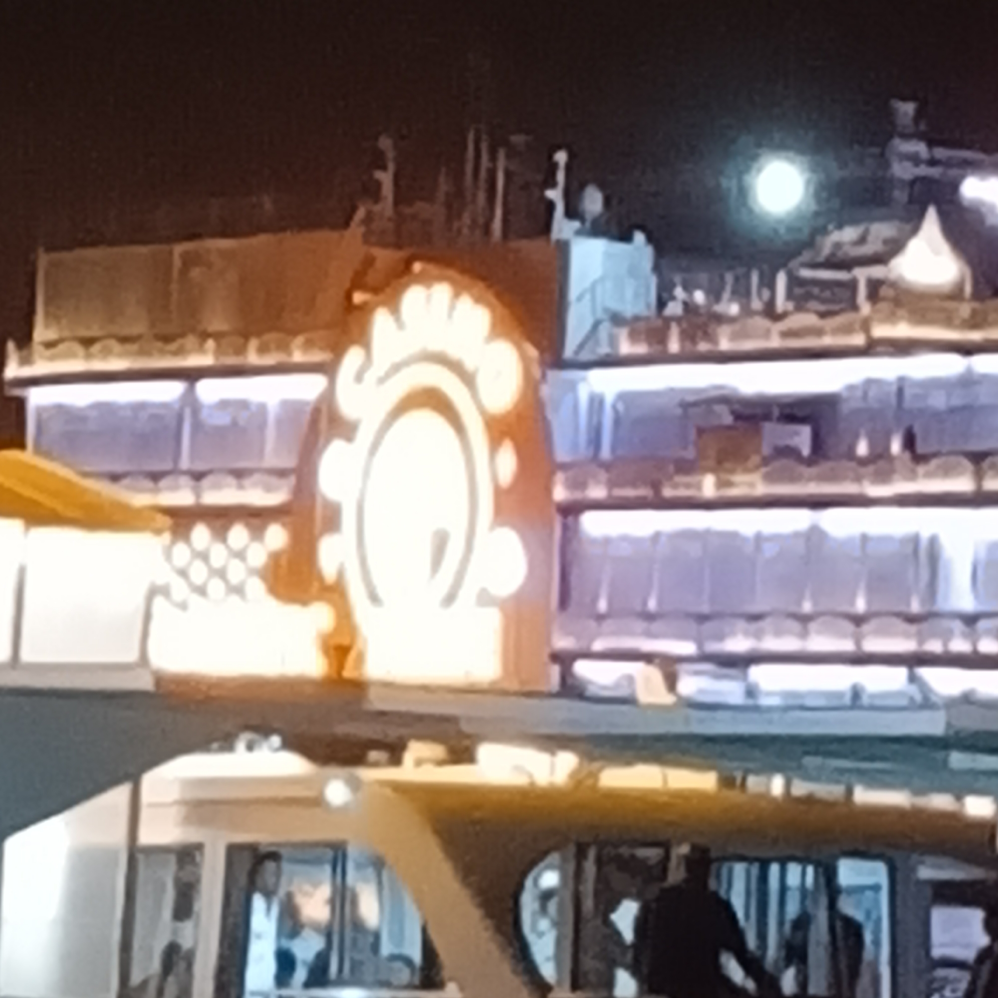 Here is a description of the image:
The image shows a building with illuminated windows and a decorative illuminated feature with a circular design. A bus or ferry is in the foreground, with several people visible through the windows. The scene appears to be at night, with a light source in the sky. The architectural style and signage are consistent with Panaji, India. The scene depicts urban activity at night.