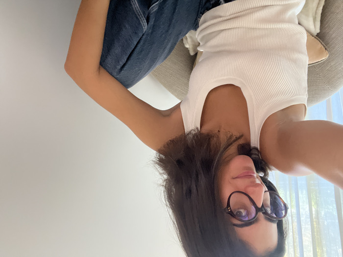 A female individual is depicted in an indoor setting located in Aldoar, Foz do Douro e Nevogilde, Portugal. The subject, positioned centrally, is seated and appears to be taking a selfie with her right arm extended, holding an unseen device. She has shoulder-length dark hair, a medium complexion, and is wearing round, tortoise-shell framed eyeglasses. Her attire consists of a white, ribbed scoop-neck tank top and dark blue denim jeans. A thin, gold-colored necklace is visible. She is seated in a light grey upholstered armchair, with a light beige decorative cushion positioned on its armrest or lap. Behind the subject, to the left, a window with sheer light-colored curtains is partially visible, indicating a source of natural light. The background to the right consists of a plain white wall. The subject's gaze is directed towards the camera.
