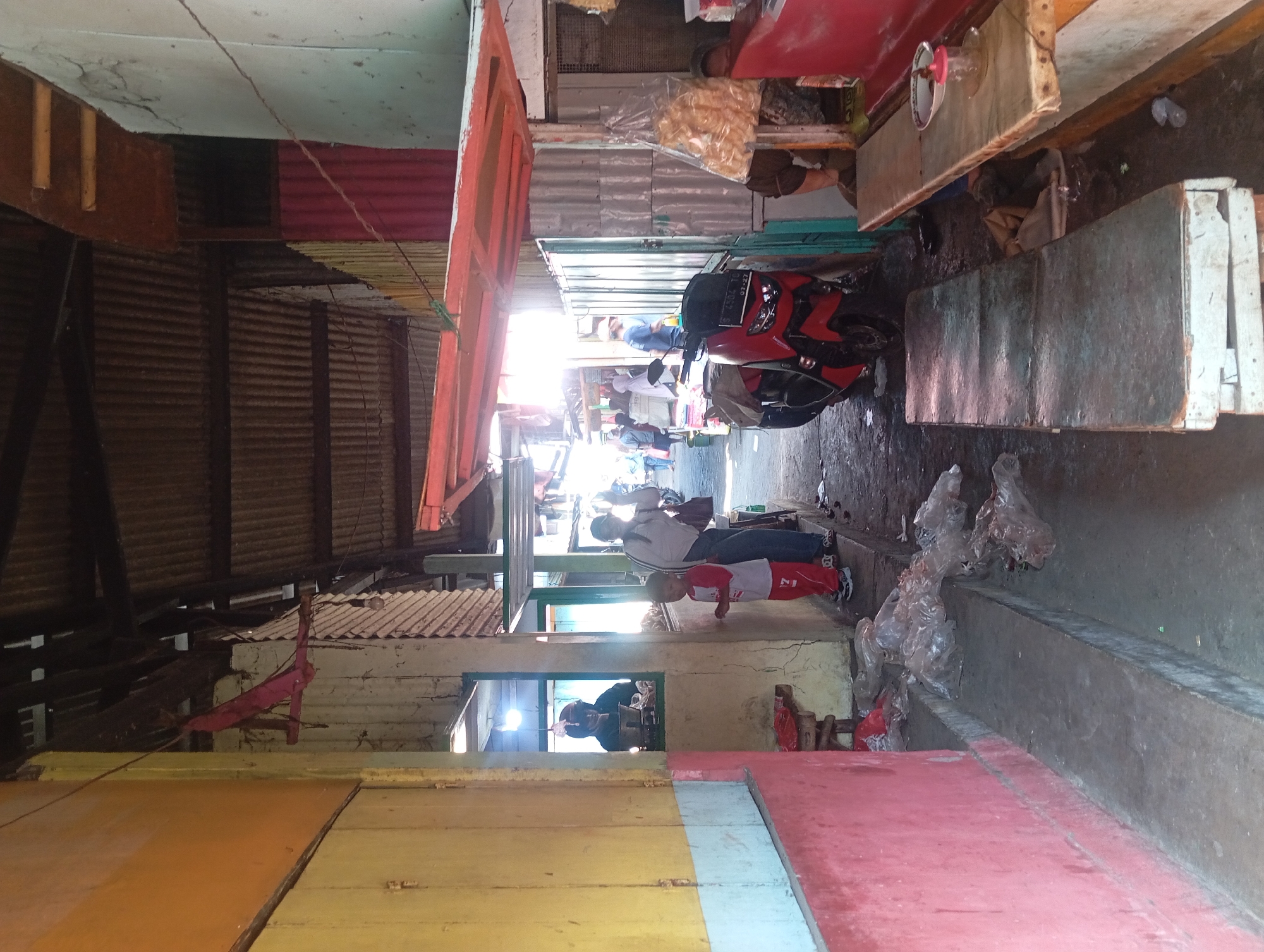 The image depicts a street scene in Kepulungan, Indonesia, likely a market area. The foreground shows a narrow walkway with a concrete bench or platform on the left and a seating area on the right. The left side of the walkway is bordered by a building with yellow and red walls. Plastic bags and debris are scattered on the ground.
In the middle ground, a red scooter is parked. To the right of the scooter, a man in a blue shirt is visible. Further back, a woman in a white headscarf and a child in a red and white outfit are standing near an opening in a building. A person wearing a helmet is visible inside this opening, possibly a vendor. The background reveals more stalls and people, suggesting a busy environment. The overhead structure appears to be covered with corrugated metal roofing supported by wooden beams. The overall atmosphere suggests an outdoor market with various vendors and shoppers.