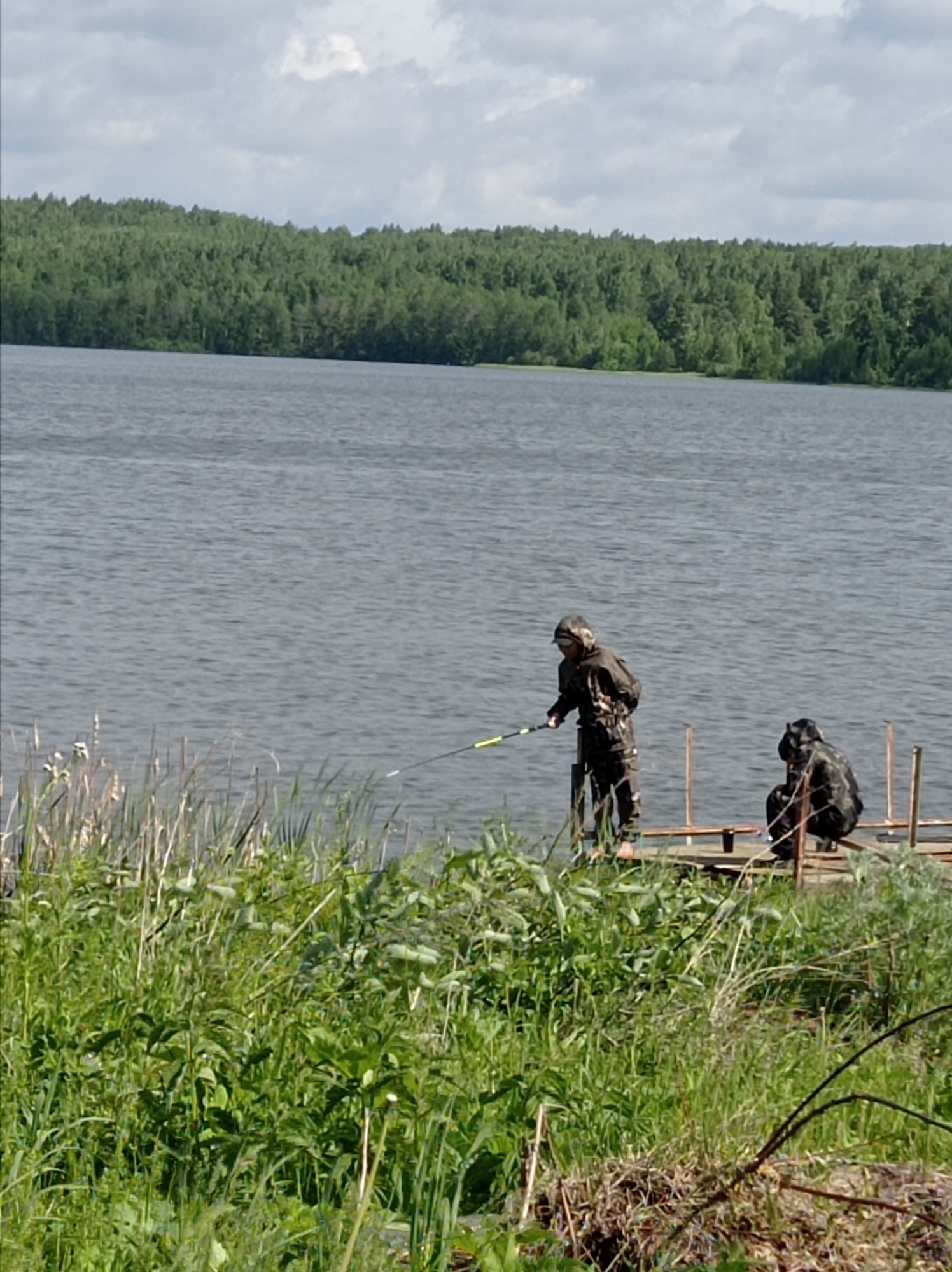 Two individuals stand on a wooden dock extending into a body of water. One person, wearing camouflage clothing, is standing and holding a fishing rod. The second person, also in camouflage, is sitting on the dock. Lush green vegetation is visible in the foreground, and a forest lines the opposite shore. The sky is overcast with clouds. This scene may be located in the city of Degtyarsk, Russia.