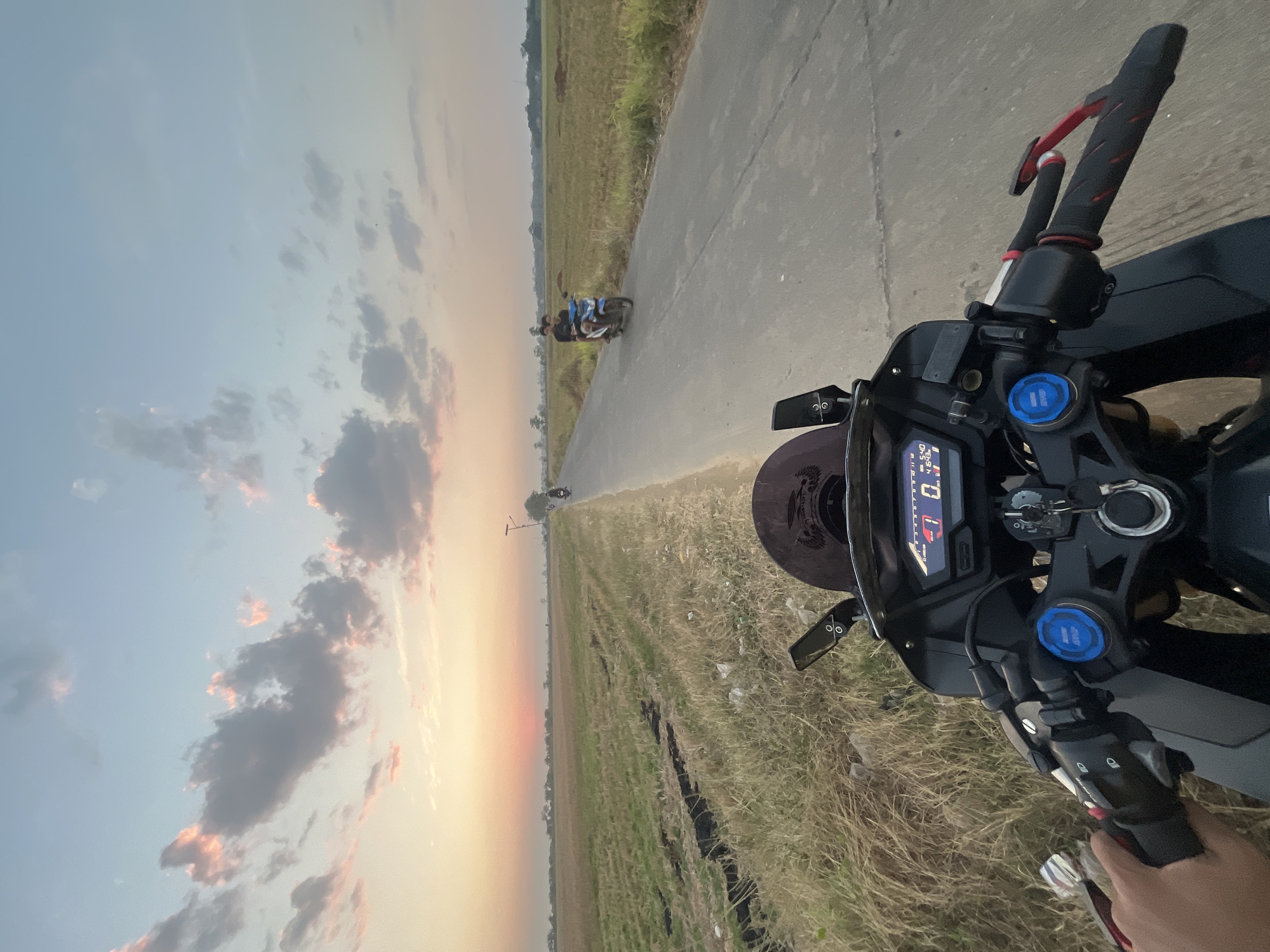 This is a first-person perspective photo taken from a motorcycle on a paved road in Pecangaan, Indonesia, during sunset. The road runs through a rural area with dry, grassy fields on either side. The sky is a gradient of soft orange and pink near the horizon, transitioning to a pale blue with scattered clouds.
In the foreground, the handlebars and digital display of the motorcycle are visible. The display shows a speed of 0 km/h and the time as 5:40. A hand is partially visible on the left handlebar.
Further down the road, two other motorcycles are present. One motorcycle, with two people riding it, is traveling in the same direction as the viewer. Another motorcycle is further ahead, with a single rider. The landscape is characterized by flat terrain with scattered trees in the distance. The overall scene suggests a peaceful evening ride through the countryside.